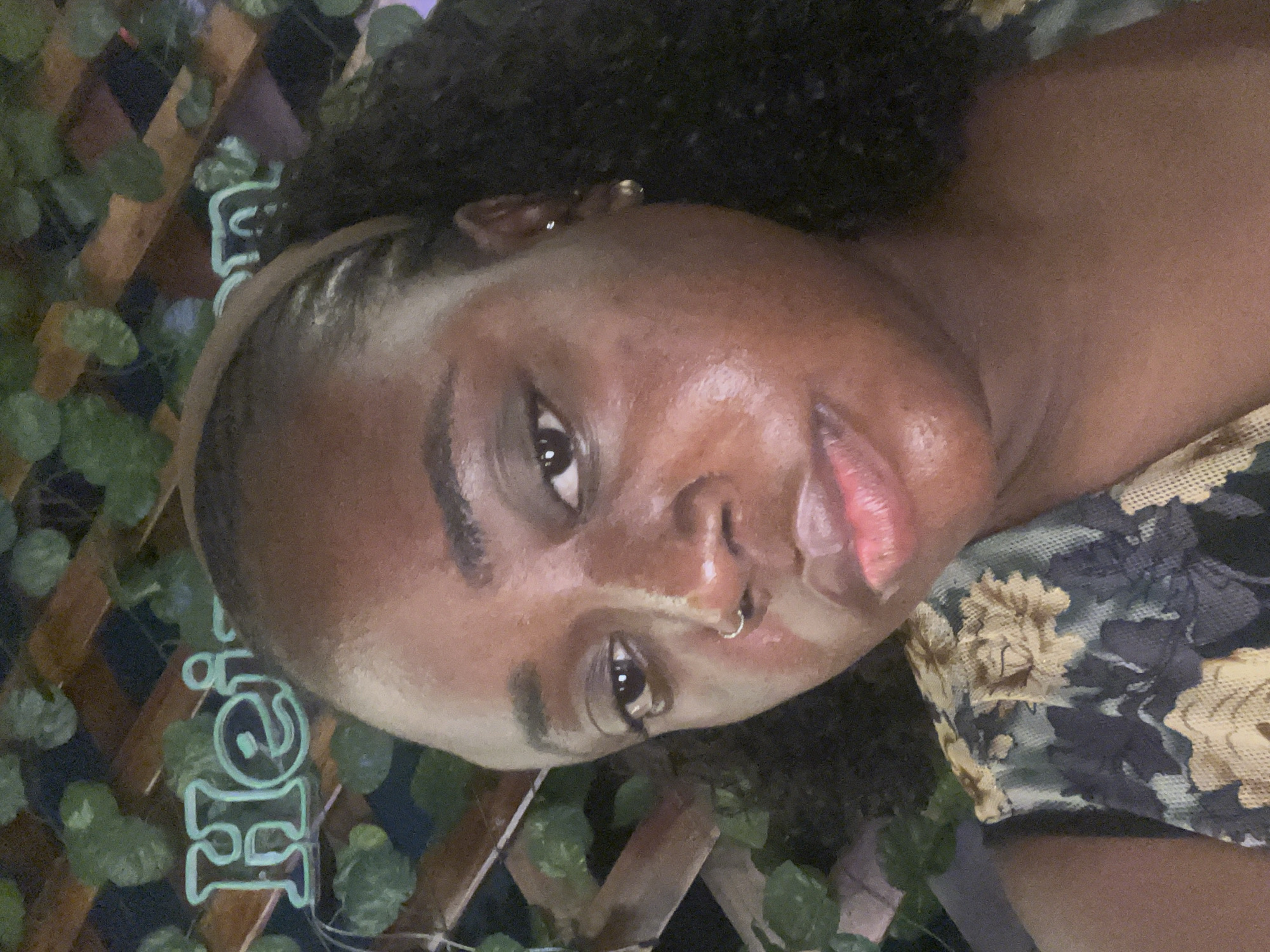 In this close-up selfie, a young Black woman with dark skin and curly hair is the central subject. She is wearing a floral top and a brown headband. Her facial features include a prominent nose ring, a small earring in her left ear, and natural makeup that highlights her lips and eyes. The background of the image is blurred, but it appears to be an outdoor setting with green foliage and a wooden lattice structure. A neon sign with the word "Heineken" in glowing green letters is visible in the upper left portion of the image. The overall lighting suggests it may be evening or nighttime. There is no indication of the specific location within Abuja, Nigeria, as the image lacks any identifiable landmarks or contextual clues specific to that city.