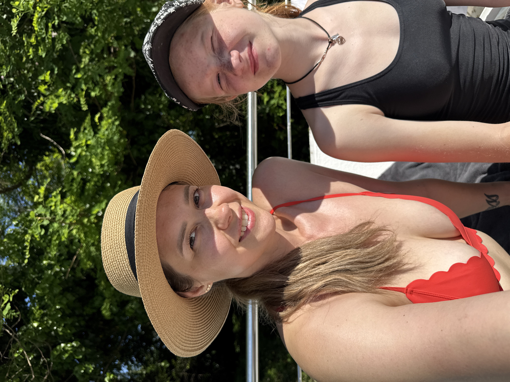 The image depicts two females, an adult and a pre-teen/teenager, outdoors in bright sunlight, positioned in the lower two-thirds of the frame. The background consists of blurred green foliage, likely trees, under a bright sky, suggesting a natural outdoor setting. A silver-colored metal railing runs horizontally behind the subjects at mid-chest level.
The adult female on the left has light brown hair pulled back in a ponytail, with strands visible over her left shoulder. She wears a light brown straw boater hat with a black band. She is smiling directly at the camera, her eyes open, and her teeth visible. Her skin appears fair and illuminated by strong sunlight, showing highlights on her forehead, nose, and cheekbones. She is wearing a red bikini top with a scalloped edge along the bustline. A small, light-colored stud earring is visible in her left ear. Her right arm is mostly cropped out of the frame.
The younger female on the right has fair skin, reddish hair, and numerous freckles on her nose and cheeks. She wears a dark-colored (likely black) baseball cap with a subtle white or light-colored pattern on its brim. She is squinting slightly, particularly her right eye, and has a faint smile directed towards the camera. She wears a black tank top and a dark cord necklace with a silver-colored charm, possibly a heart or lock design. Her left arm is mostly cropped out of the frame.
Both individuals are positioned side-by-side, facing forward. The lighting suggests a clear, sunny day. The image provides no specific visual indicators to confirm a precise geographic location such as Nebug, Russia.
