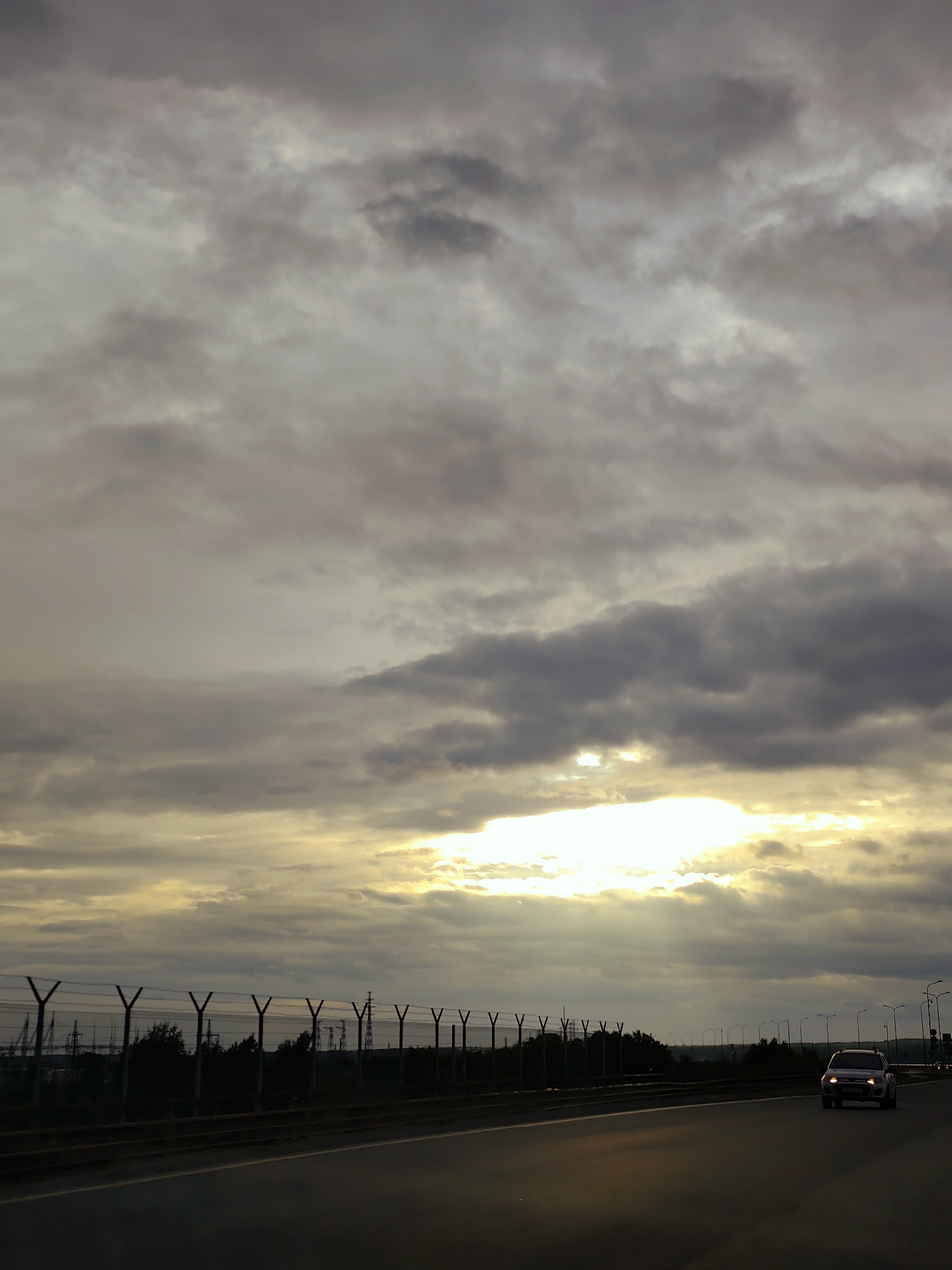 The image captures a multi-lane asphalt highway under a heavily clouded sky during either sunrise or sunset. The upper two-thirds of the frame are filled with dense, grey stratocumulus clouds. A pronounced break in the cloud cover in the lower central sky reveals an intense burst of golden-yellow light, illuminating the undersides of surrounding clouds and casting visible light shafts downwards.
The highway occupies the lower third of the image, extending from the foreground into the distance. On the left side of the road, a metal security fence with distinctive Y-shaped posts topped with multiple strands of barbed wire runs horizontally. Behind this fence, a dark band of trees and shrubs is visible, followed by numerous tall, silhouetted power transmission towers (pylons) against the brighter sky, indicating the presence of a significant electrical infrastructure or industrial complex. On the right side of the highway, a series of streetlights on tall poles recedes into the background.
A single white sport utility vehicle (SUV) with its headlights illuminated is present in the rightmost lane, traveling away from the viewer. No other vehicles or people are visibly present.
The scene's industrial elements, particularly the extensive power pylons and security fencing, are consistent with areas surrounding major energy infrastructure. In the city of Chaikovsky, Russia, such an environment could be found on highways in proximity to the Votkinsk Hydroelectric Power Plant on the Kama River or other industrial zones within the city's operational periphery.
