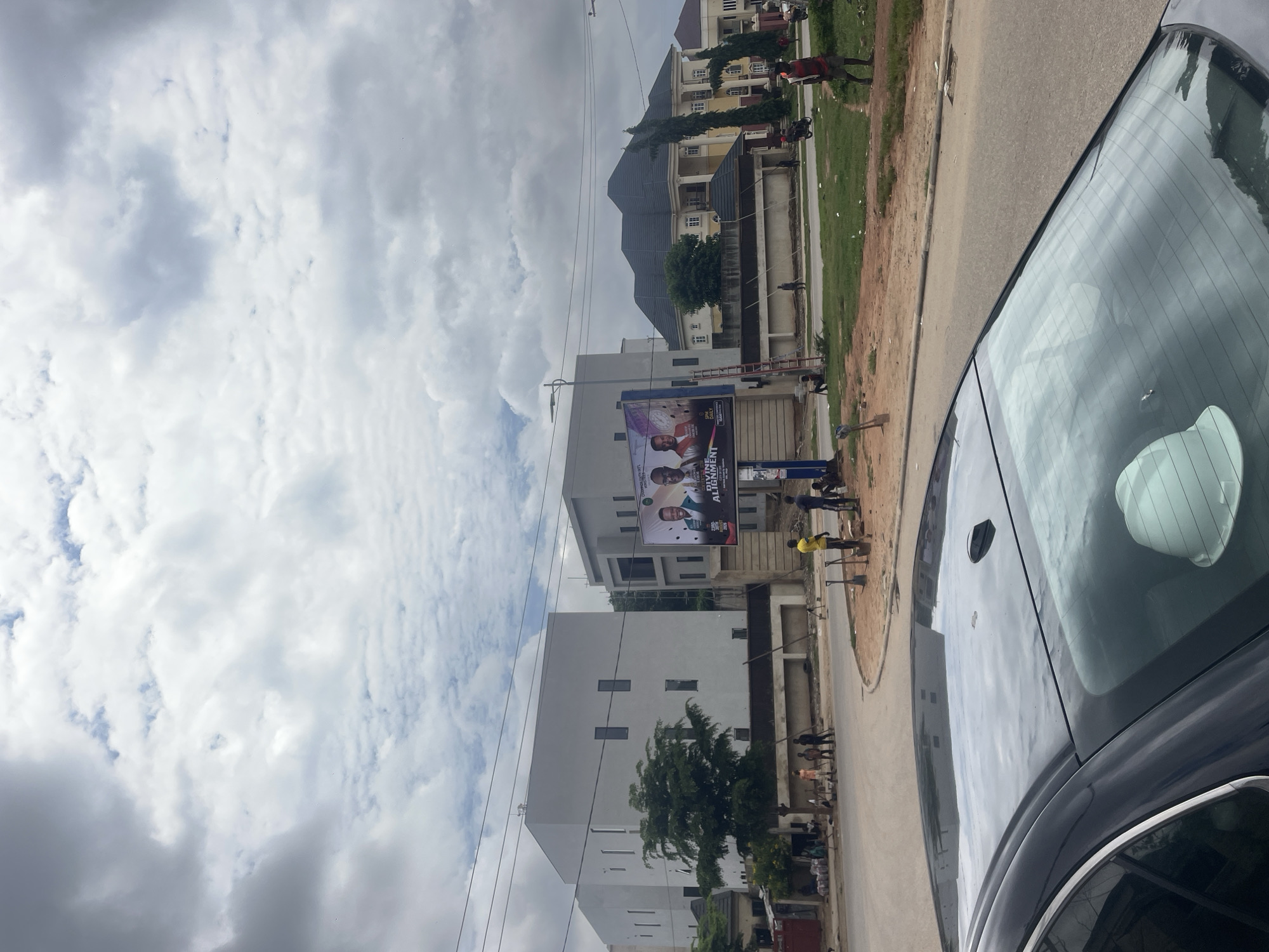 A wide-angle, vertical shot captures a street scene in Guzape, Nigeria, under a cloudy sky. In the foreground, the reflective black roof of a car is visible, with a white hard hat on its rear window. A paved road curves to the left and extends into the background.
Across the road, a billboard displays images of two men in suits, with text that appears to be in English. To the left of the billboard are modern, minimalist white buildings with rectangular windows. Further back, a large, traditional yellow house with a dark, multi-pitched roof stands prominently.
Several individuals are scattered throughout the scene. Some appear to be workers, with tools like shovels visible on the ground. One person is standing near a ladder next to the billboard. A motorcycle is also seen in the mid-ground, with a rider and passenger. The environment is a mix of urban development and open, grassy areas.