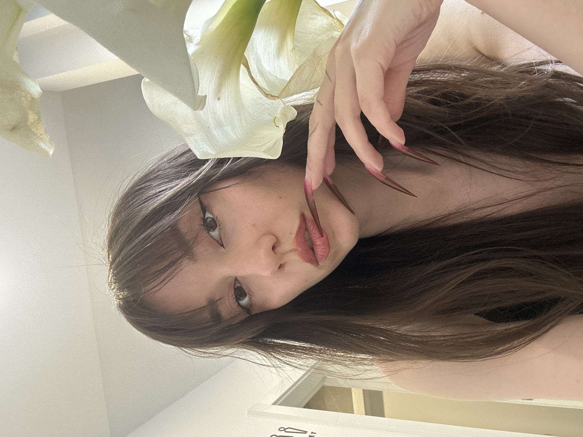 A young woman poses with a calla lily. The woman has long brown hair and long, pointed, red painted nails. She is looking at the camera with her lips pursed. The scene includes a partial view of an interior with a white door and white wall. The location is not discernible as Vracar Urban Municipality, Serbia.