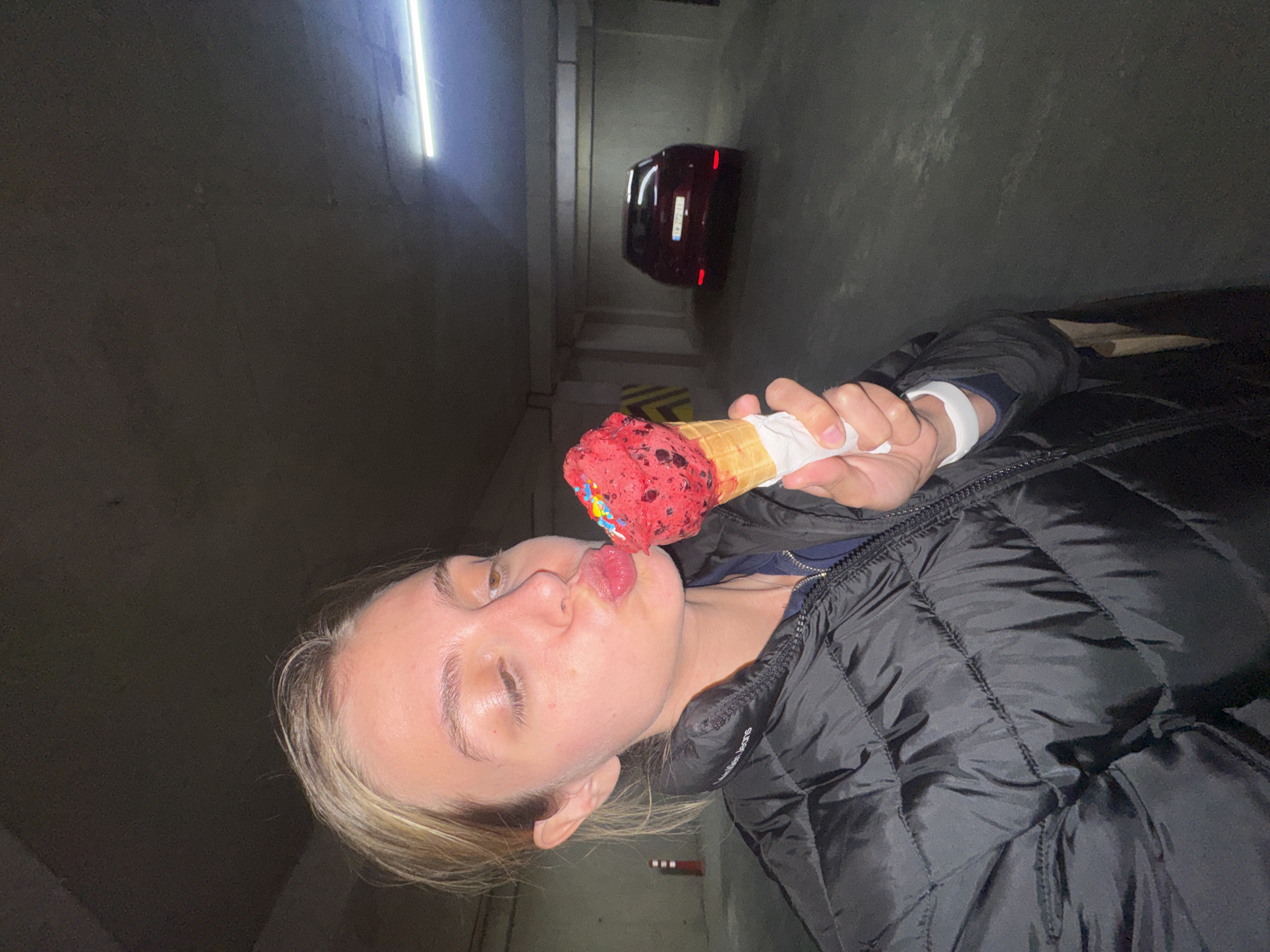 A woman with blond hair and fair skin stands in an underground parking garage and eats an ice cream cone with red ice cream and colorful sprinkles. She is wearing a black puffer jacket. Behind her, a red car is parked in the garage, which is lit by fluorescent lighting. The city is unknown.