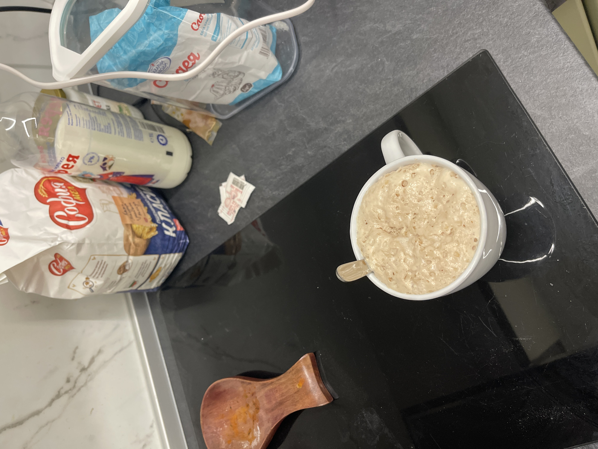 The image shows a kitchen countertop, likely in Sofia, Bulgaria due to Cyrillic text on food packaging. A white mug filled with a bubbly, light brown mixture rests on a black induction cooktop. A small, clear plastic spoon sits in the mug. Adjacent to the cooktop, there's a wooden spoon rest with orange residue. Scattered nearby are food ingredients: a bag of flour labeled "Rodina," a bottle of milk, and a package of what appears to be sugar. A white plastic container with blue and white packaging is also visible. The countertop surface is a speckled gray, and a section of white marble countertop is visible in the lower-left corner.