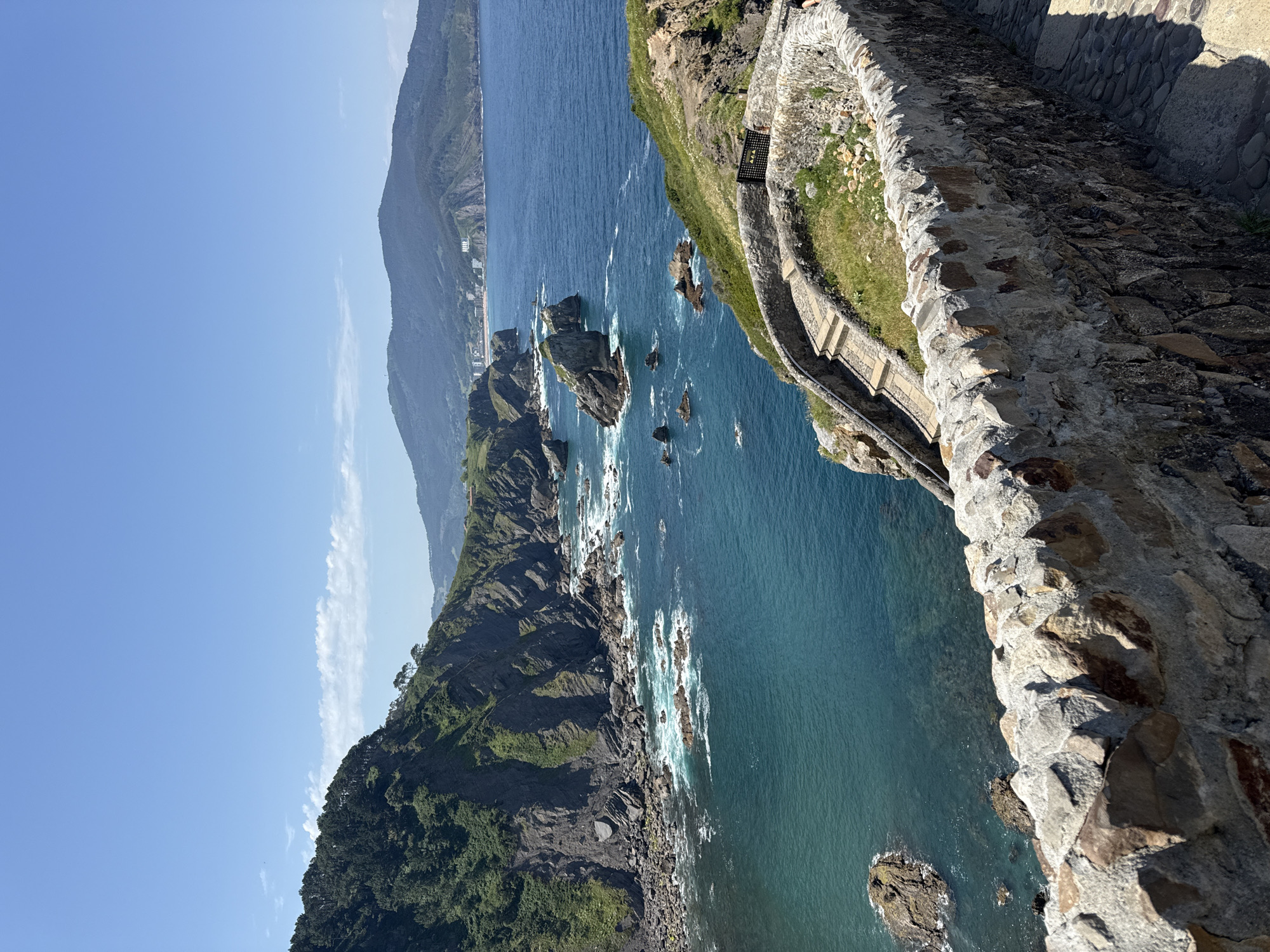 This image captures a scenic view of the coastline near Bakio, Spain. In the foreground, a rugged stone wall, constructed from various grey and brown stones, curves along a path. Beyond the wall, a stone staircase descends along the grassy cliffside. The sea, a vibrant turquoise color, crashes against rocky outcrops and a steep, green-covered cliff face on the left. In the distance, a range of hills forms the backdrop, with buildings visible on the slopes and a faint haze in the sky. The sun is shining, casting shadows on the path and highlighting the textures of the stone and landscape. The scene appears peaceful and natural, showcasing the beauty of the Basque Country's coastline.