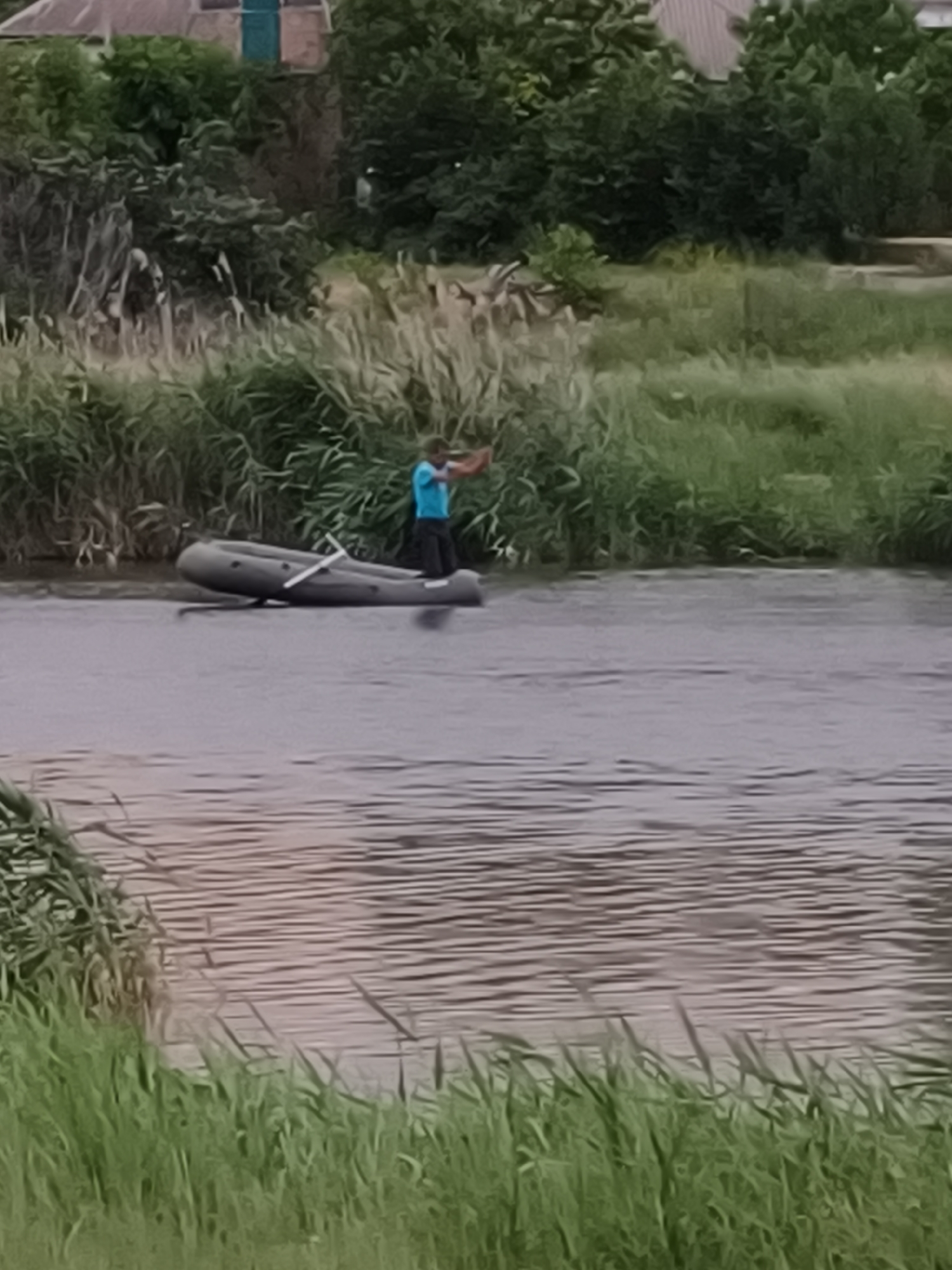 An individual in a blue t-shirt and dark pants stands in a gray inflatable boat on a body of water. The boat has a white oar resting inside it. The individual is holding a dark object in their right hand, and their left arm is bent at the elbow. The water is a muted gray color with ripples. On the far bank, dense green vegetation, including reeds and taller grasses, lines the water's edge. In the background, beyond the vegetation, there are glimpses of buildings with gray roofs and green doors, surrounded by trees. The scene appears to be outdoors, possibly in a rural or suburban area on the outskirts of Kryvyi Rih, Ukraine, given the presence of the water, vegetation, and residential structures.