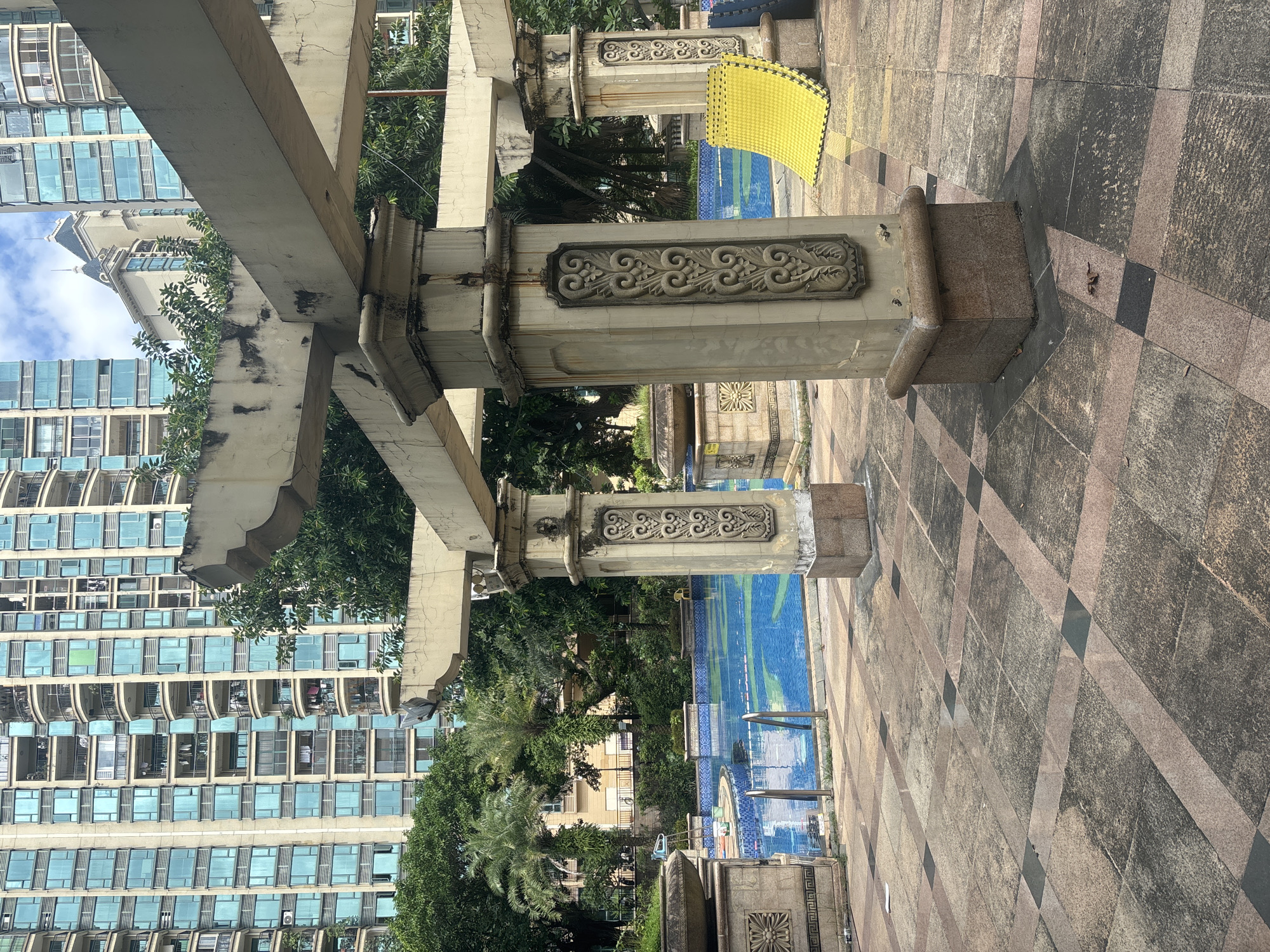 The image shows an outdoor swimming pool area in front of a residential building in Nanhai District, China. The pool is blue and tiled, with a few lounge chairs and pool accessories visible. A covered patio structure with stone pillars and decorative carvings stands in the foreground, casting shadows on the patterned stone pavement. Lush green trees surround the pool area, and a tall, modern residential building with numerous balconies and blue-tinted windows looms in the background. The sky is partly cloudy. The scene depicts a leisure space within a densely populated urban environment.
