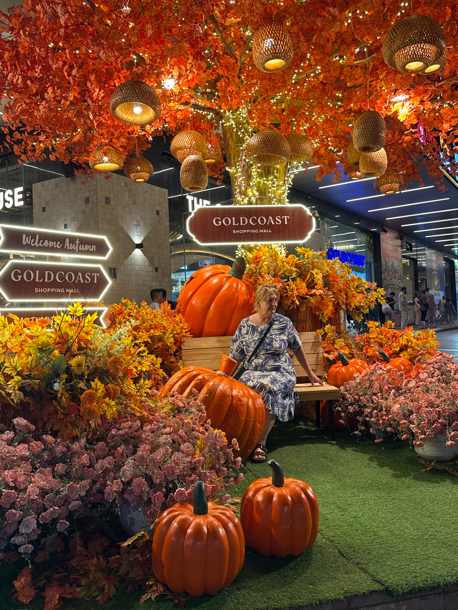 The image depicts an outdoor autumnal display, likely a seasonal decoration in a public space, potentially a shopping mall or plaza in Nha Trang, Vietnam. A woman, dressed in a blue and white patterned dress and sandals, is seated on a wooden bench. She is looking to her left, away from the camera.
In front of the bench, there is a collection of large, artificial pumpkins and an abundance of artificial autumn foliage in shades of orange, yellow, and red. There are also clusters of small pink flowers, and some dried leaves scattered on the artificial grass. A large, decorative pumpkin is prominently placed to the left of the bench.
Above the scene, a canopy of artificial autumn leaves is illuminated by numerous small lights and several woven pendant lamps. In the background, a building with a stone facade is visible, along with signage indicating "GOLDCOAST SHOPPING MALL" and "Welcome Autumn." The overall impression is a festive and decorative arrangement for the autumn season. The presence of "GOLDCOAST SHOPPING MALL" strongly suggests this location is within or associated with that establishment.