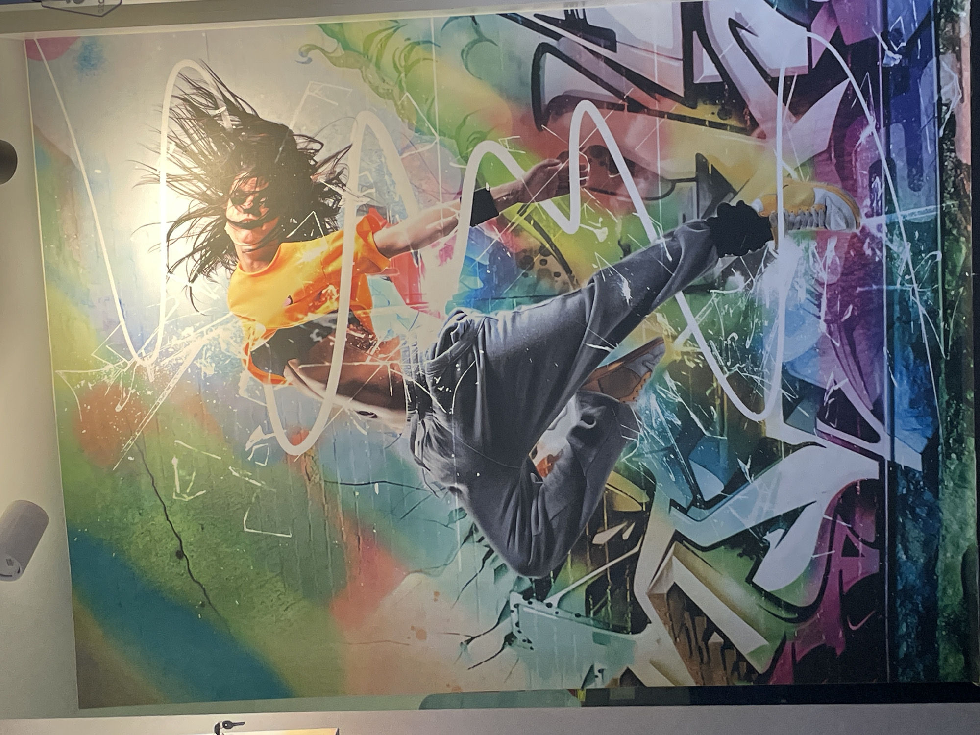 The image shows a wall mural. The mural depicts a person in mid-air, executing a dance move. The person wears an orange shirt and grey pants. Their hair is in motion, suggesting dynamic movement. The background features a graffiti style design with vibrant colors and bold shapes. The mural is found in the city of Porlamar, Venezuela.