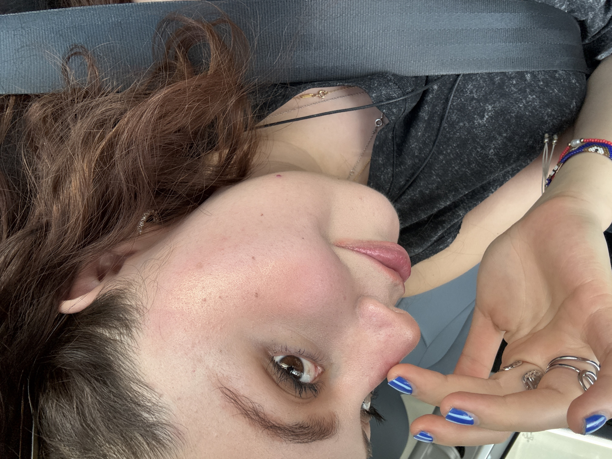 A close-up, angled portrait shows a person with light skin, brown hair, and brown eyes. The person is wearing a grey speckled shirt and a seatbelt. They have manicured fingernails painted blue with white stripes. Multiple rings are visible on their fingers. The person's head is tilted, and their gaze is directed upwards. Their lips appear slightly parted. Three necklaces are visible around the person's neck. The location and specific city or environment in Mexico City are not discernible from the provided image.