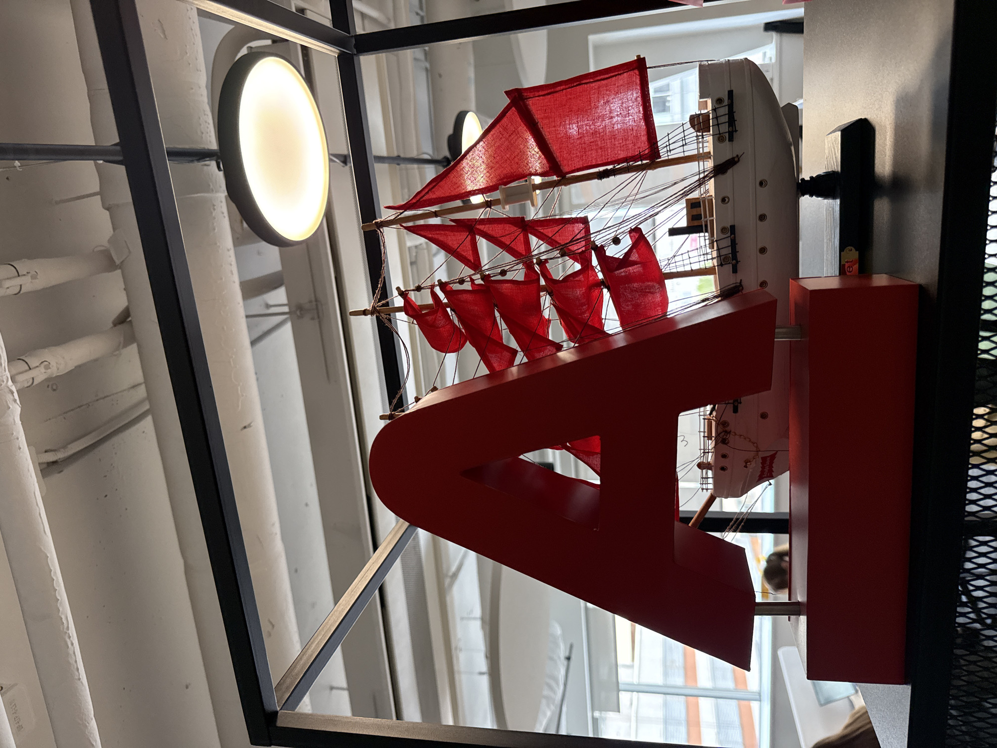 The image shows a red model sailing ship with its red sails unfurled, suspended within a black metal frame. The ship is attached to a red structure resembling a letter, possibly "A" or a stylized emblem. The backdrop features a wall with vertical, ribbed patterns. In the background, a circular light fixture emits a soft glow. The location appears to be indoors, based on the lighting and structural elements. The city of Unknown, Unknown is not identifiable.