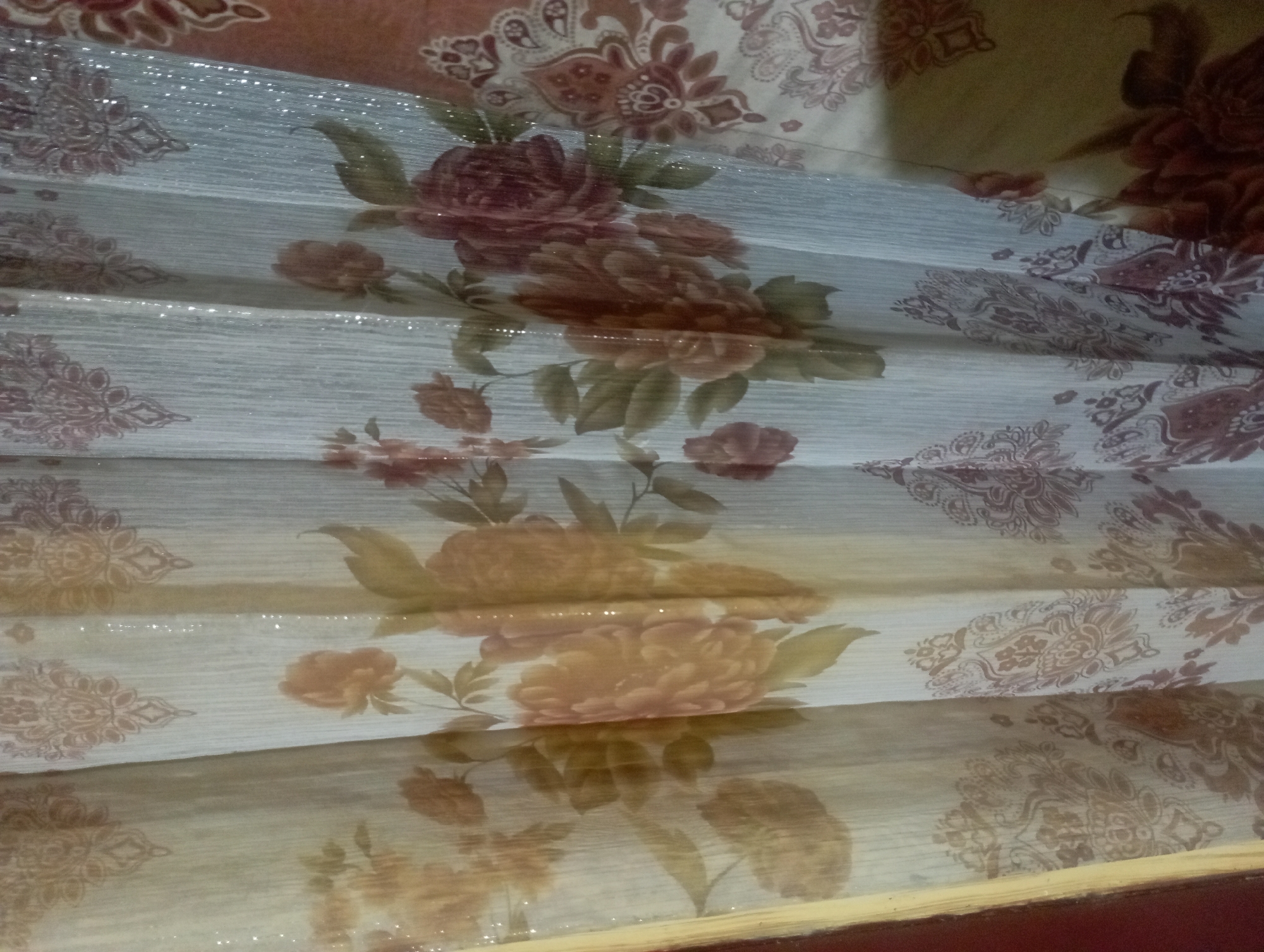 A close-up, eye-level, static medium shot captures a section of a curtain with a floral pattern. The curtain is a light beige with a textured vertical weave, and it is adorned with a repeating pattern of large, stylized flowers in shades of peach, orange, and deep red, as well as smaller floral motifs and ornate paisley designs in brown and maroon. Shimmering, glitter-like particles are embedded within the fabric, catching the light and creating a speckled effect throughout the material. The curtain is draped, with several vertical folds creating a textured, layered appearance. The lighting is somewhat dim, with the glittery elements being the brightest points in the image. The scene appears to be indoors, likely a domestic setting. There are no people or specific actions depicted. The location is a residential area in Maos Kidul, Indonesia.