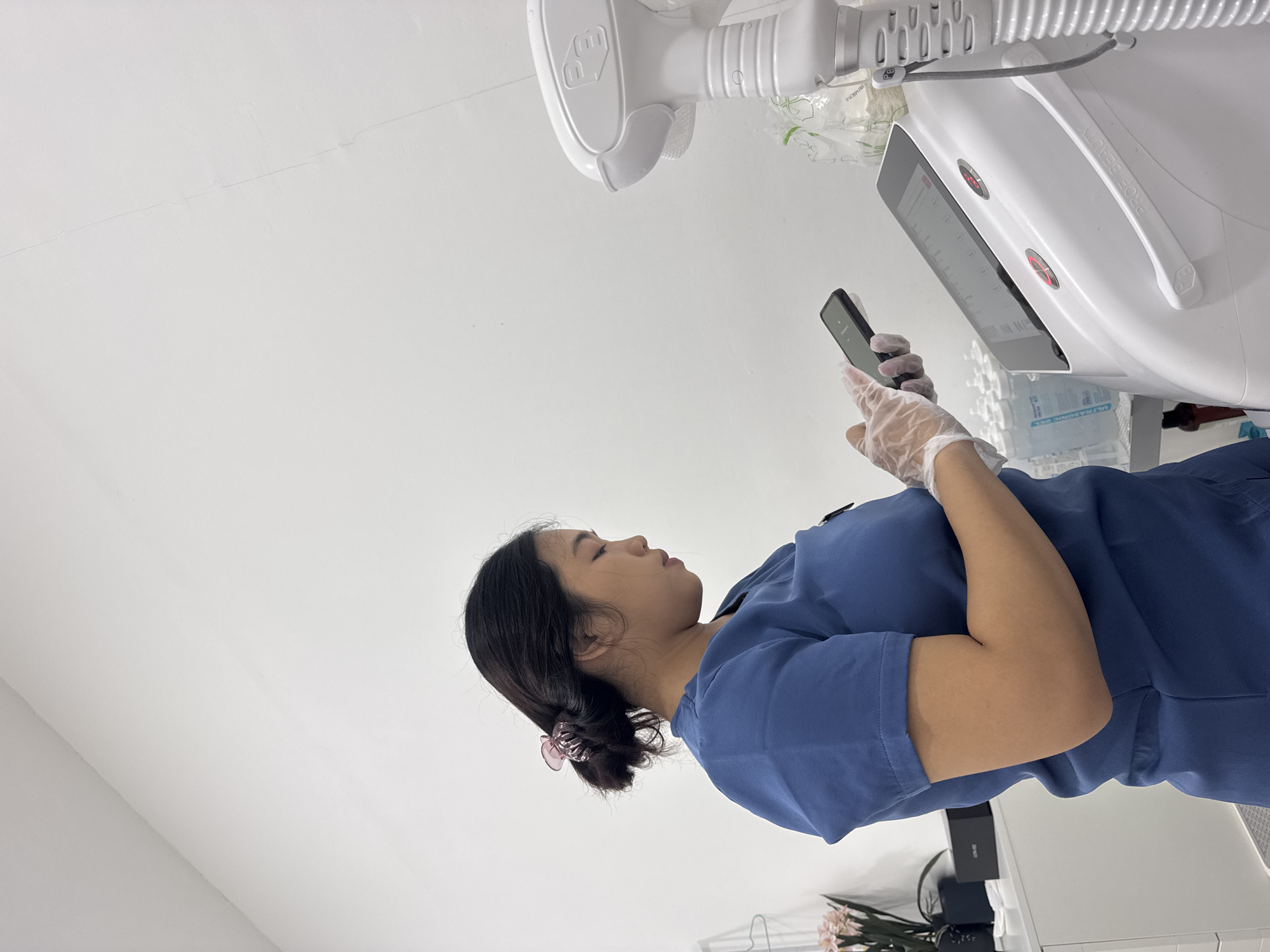 An Asian woman, wearing a blue scrub top and clear disposable gloves, stands indoors next to a large white medical or aesthetic machine. Her dark hair is pulled back with a pink claw clip, and she is focused on a black smartphone she holds in her gloved hands. Her expression is neutral, suggesting concentration on her phone.
The setting appears to be a professional environment, such as a clinic, spa, or beauty salon, with plain white walls. The prominent white machine features a digital screen displaying an interface, handles, and tubing. Text visible on one of the machine's handles reads "PROF BEAUTY," and a "P_D" or similar logo is also visible. Numbers "0.0.0.0.0" are seen on an upper part of the machine. In the background, several bottles are stacked, with "ULTRASONIC GEL" clearly legible on some labels. Faux pink and white flowers are arranged on a surface to the woman's left, and a black box is also present. The scene is brightly lit, indicative of an indoor daytime setting. The overall context suggests a professional taking a moment to check her phone while on duty in Kuta Selatan, Indonesia.