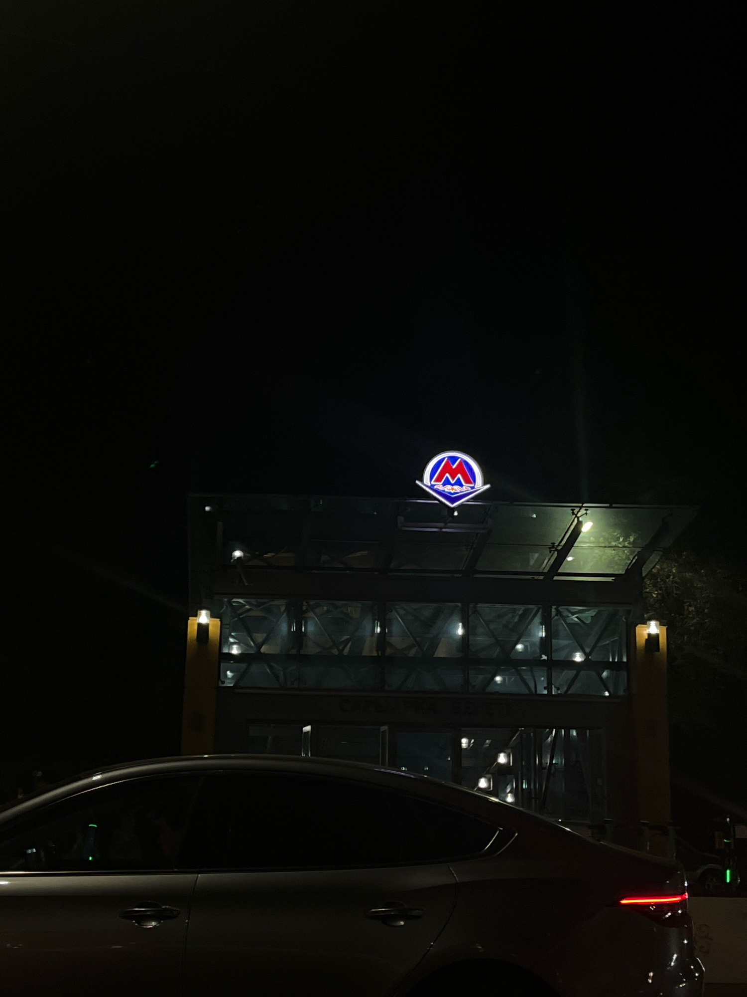 The image shows a subway entrance at night in Almaty, Kazakhstan. A silver sedan is parked in front of the entrance. The entrance is illuminated by a lit metro sign, which is red and blue with a white "M" symbol. The building has glass walls and a glass roof. The street appears to be dark and empty. The metro sign appears to be the logo for the Almaty Metro.