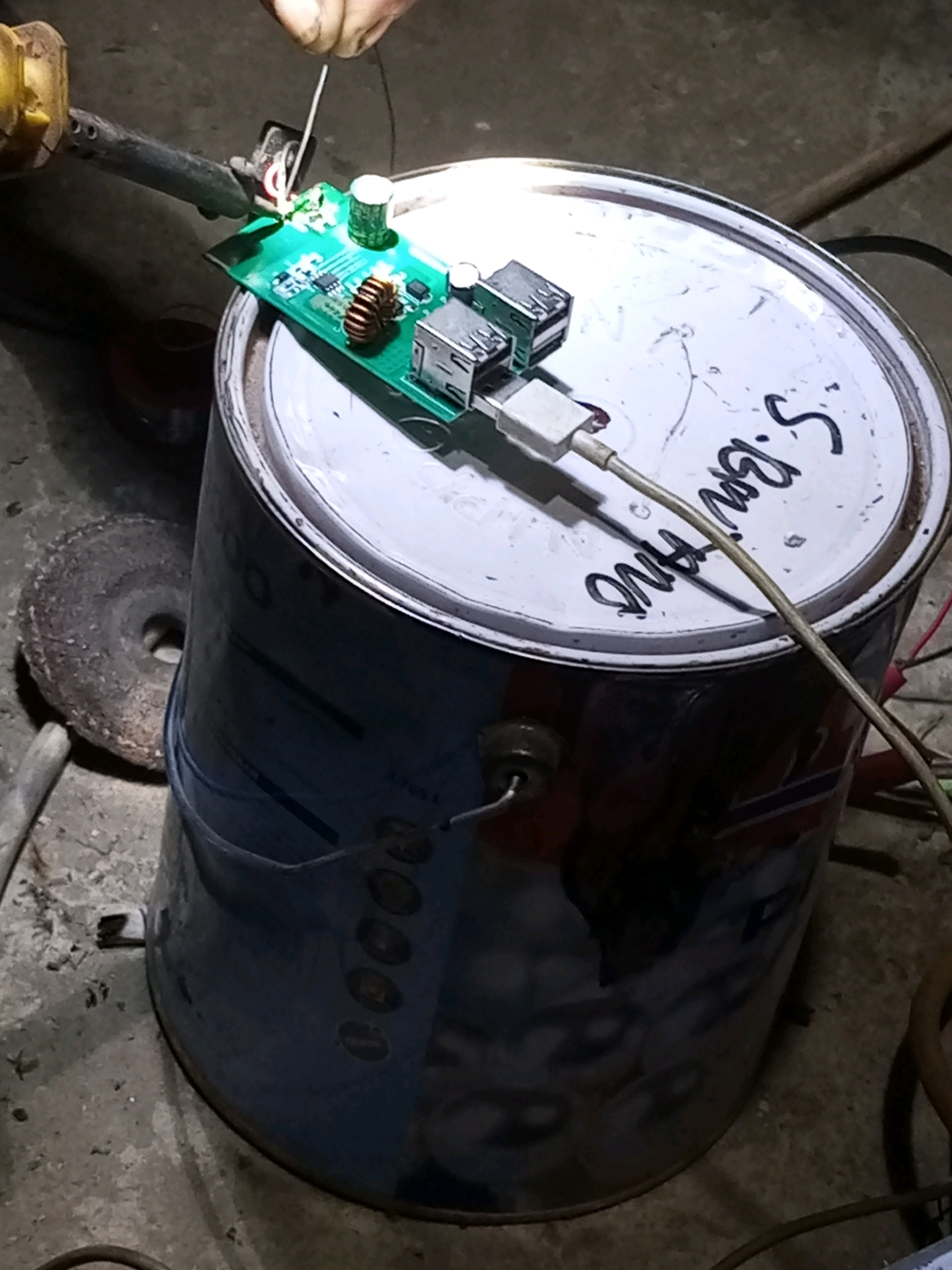 The image depicts a close-up of a DIY electronic project. A person's hands are visible, holding a soldering iron and a thin white wire, soldering a circuit board. The circuit board has several components, including two USB ports, a large capacitor, and a coil. The circuit board is positioned on top of a white-lidded metal container, possibly a paint can, which has handwritten text on its lid. A USB cable is plugged into one of the ports on the circuit board. The environment appears to be an outdoor or workshop setting, with a concrete or dirt floor visible, along with some tools, including a grinding wheel. There is no discernible information in the image that would suggest the location as Keling Panggau, Indonesia.