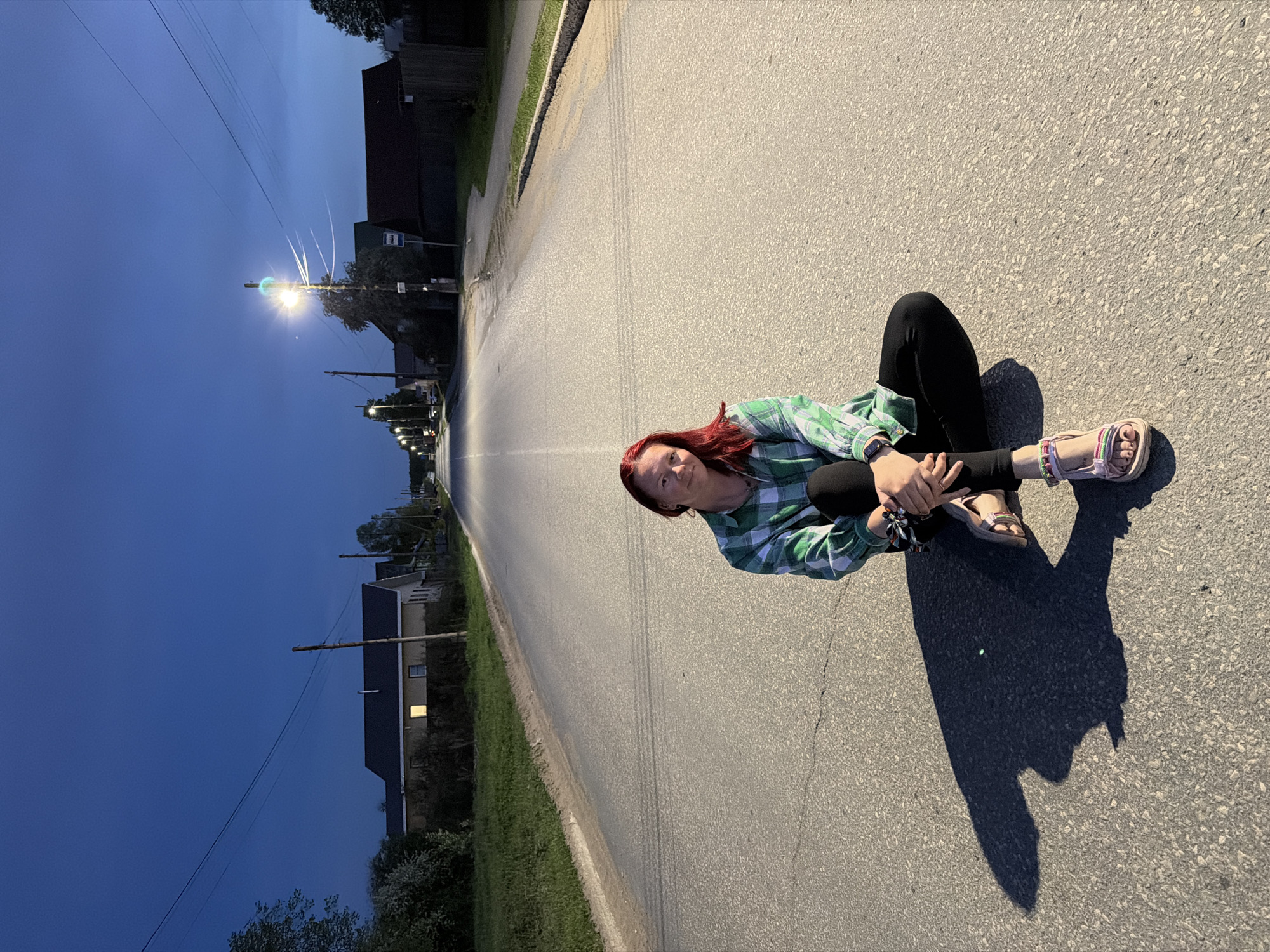A woman sits cross-legged on an asphalt road in Chaikovsky, Russia. She wears a green and white plaid shirt, black leggings, and sandals. Her hair is red. The road stretches into the distance toward a streetlight, with buildings and trees lining either side. The sky is a deep blue.