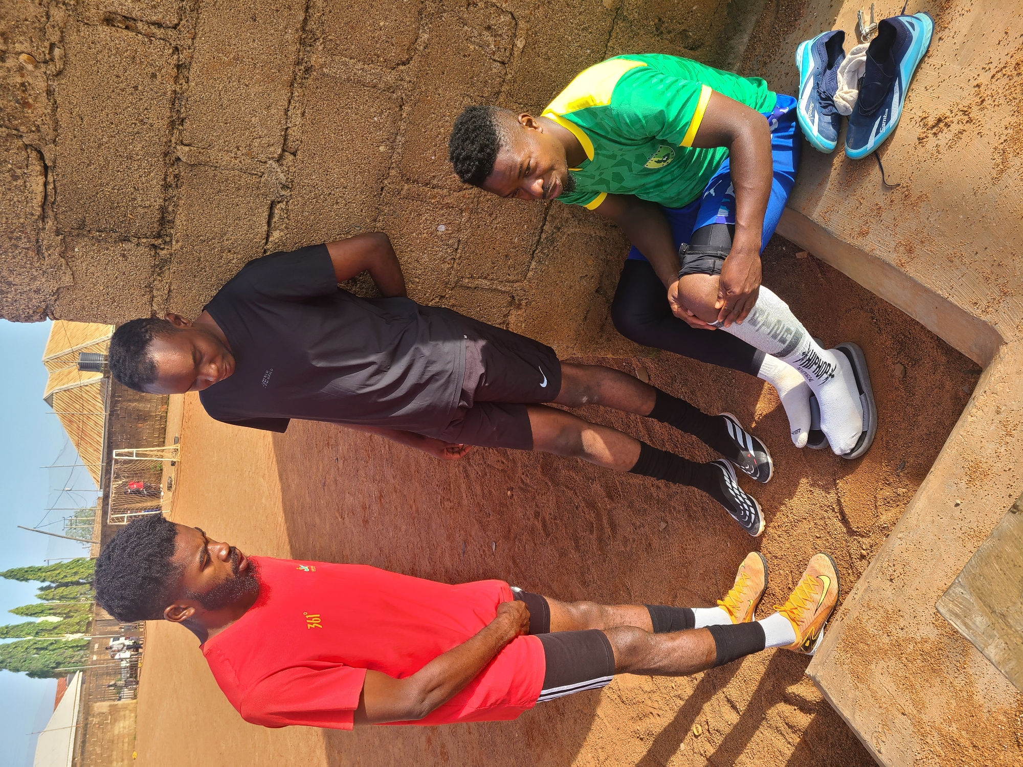 In Barakin Lerawi, Nigeria, three young men stand and sit in the mid-afternoon sun on a dusty, orange-brown soccer field. Two men are standing and one is sitting. The man on the left, wearing a bright red t-shirt with white lettering, and black shorts with white stripes, stands with his right hand resting on his left hip, looking down. Beside him, leaning against a rough, brown brick wall, a man in a black t-shirt and black shorts also looks down. To the right, a third man sits on the ground, his left leg bent at the knee and his hand resting on it. He wears a green and yellow soccer jersey and black leggings. Various soccer cleats and shoes are scattered on the ground near the seated man. In the background, a soccer goal and net are visible, along with other people and structures, suggesting a community sports area.