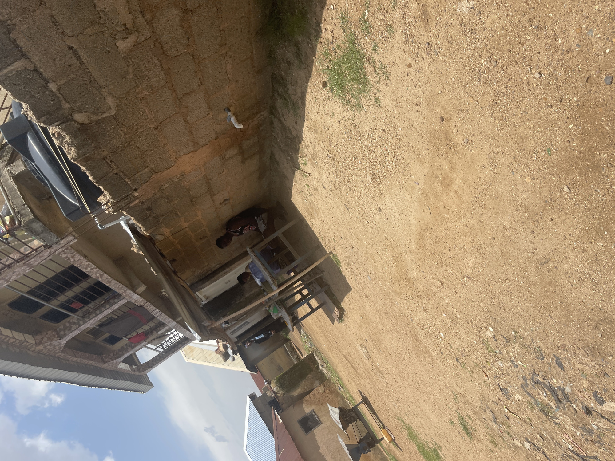 The image depicts an outdoor scene on a bright, sunny day, likely in Jos, Nigeria. The foreground is dominated by a dusty, reddish-brown ground, scattered with small stones, pebbles, and sparse patches of dry grass. In the midground, there is a cluster of low-rise buildings. One building, to the left, has a corrugated metal roof and a simple square window. Adjacent to it, a more complex, multi-story structure stands, featuring a textured facade, balconies with railings, and several windows. A large black water tank is visible on the roof of one of the buildings.
In front of the textured building, a simple setup with two small tables and benches is visible, suggesting a small outdoor seating area or a makeshift shop. Two individuals are present: one person is seated at one of the tables, appearing to be engaged with something on the table. Another person is standing further back, near the entrance of a building. A wall constructed of concrete blocks runs along the right side of the image, showing signs of wear and discoloration. A white pipe protrudes from this wall, with some water staining evident below it. The overall impression is one of a humble, possibly developing urban or semi-urban environment.