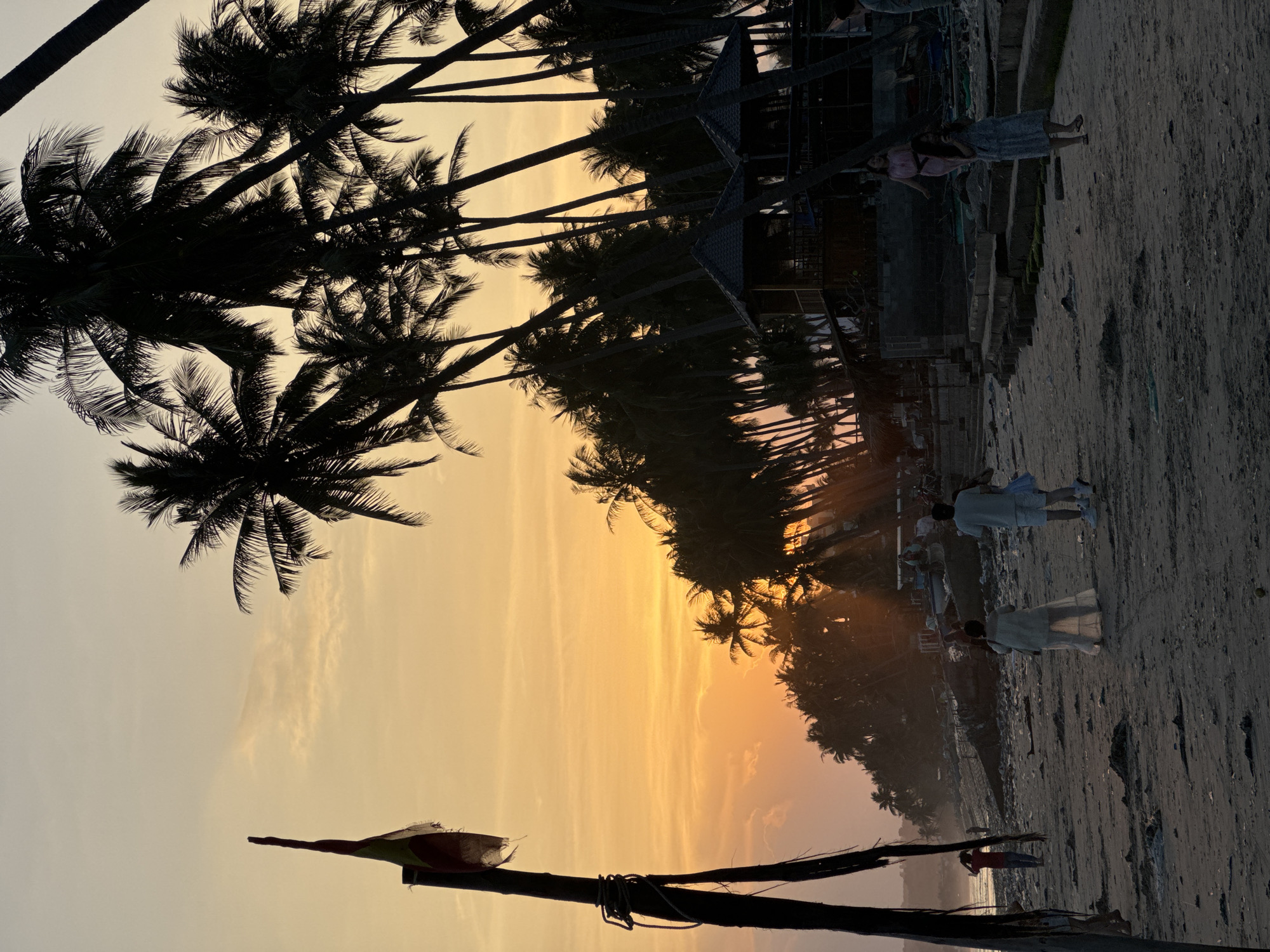 The image depicts a beach scene at sunset in Mũi Né, Vietnam. A tall wooden pole with a tattered flag is positioned on the left side of the frame. A line of silhouetted palm trees stretches across the background, with two thatched-roof huts visible among them. Several people are scattered across the sandy beach. In the foreground, two individuals, one in a light-colored outfit and the other in a white shirt and shorts, are walking. Further back, a person in a white dress is standing, and another individual in a pink dress is walking. A person in a red shirt is also visible in the distance. The sky is filled with warm orange and yellow hues, indicating the setting sun, with rays of light piercing through the palm trees. The beach appears somewhat littered with debris.