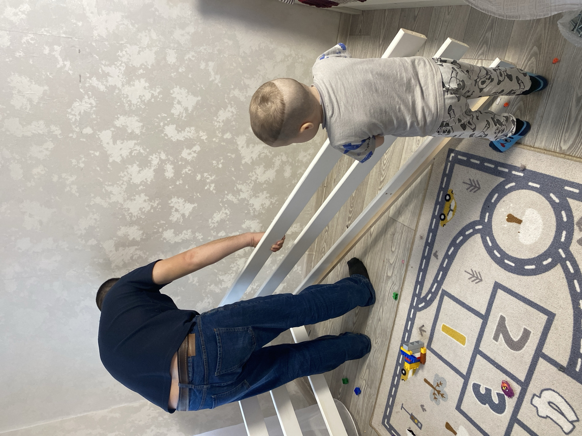 A top-down shot shows a man and a young child next to a white, partially assembled structure, possibly a children's play structure, on a wood-look floor. The man, wearing a dark blue t-shirt and blue jeans, is bent over, holding onto the structure. The child, in a gray shirt and patterned pajama pants with blue socks, is standing on the other side of the structure. To the right, a rug with a road and hopscotch design is visible, along with small toys. The background wall has a mottled gray and white pattern. The location is likely indoors and residential; there's no indication of it being in Sochi, Russia.