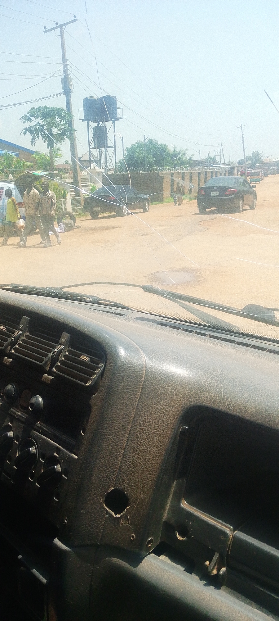 The image is taken from the interior of a vehicle, looking out through a cracked windshield. The view outside shows a dusty road in a semi-urban or rural area. Several people are visible, some appearing to be in uniform. There are cars parked and driving on the road, along with a water tower and power lines. The dashboard of the vehicle is in the foreground, showing wear and tear.