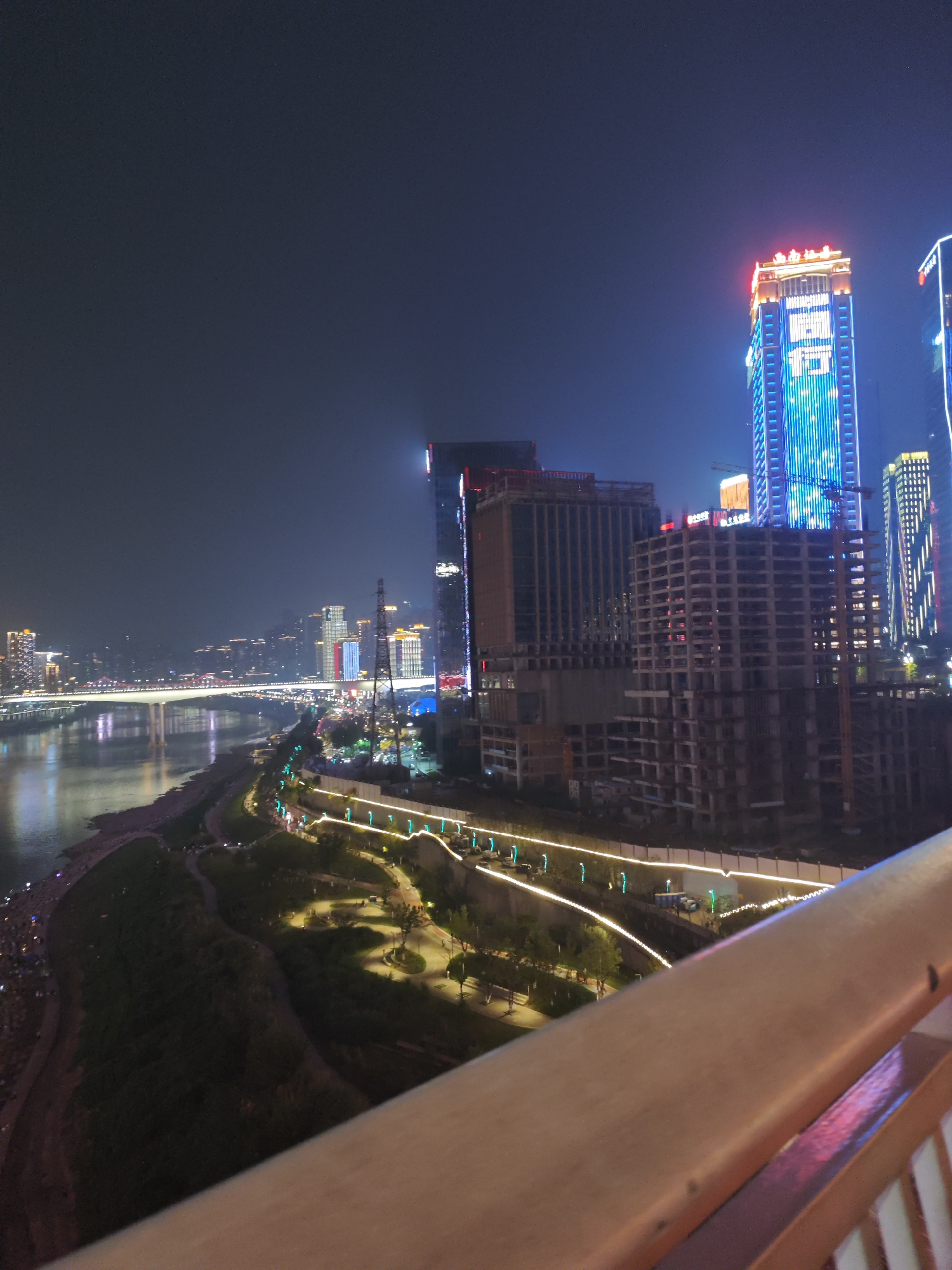 The image depicts a nighttime cityscape of Yuzhong District, China, showing a river with a bridge in the background and various lit buildings, including a tall structure with blue and red illumination. The foreground includes a landscaped area with illuminated pathways and vegetation. A portion of a railing is visible in the bottom right corner of the image. Construction sites and buildings in various stages of completion are also apparent.