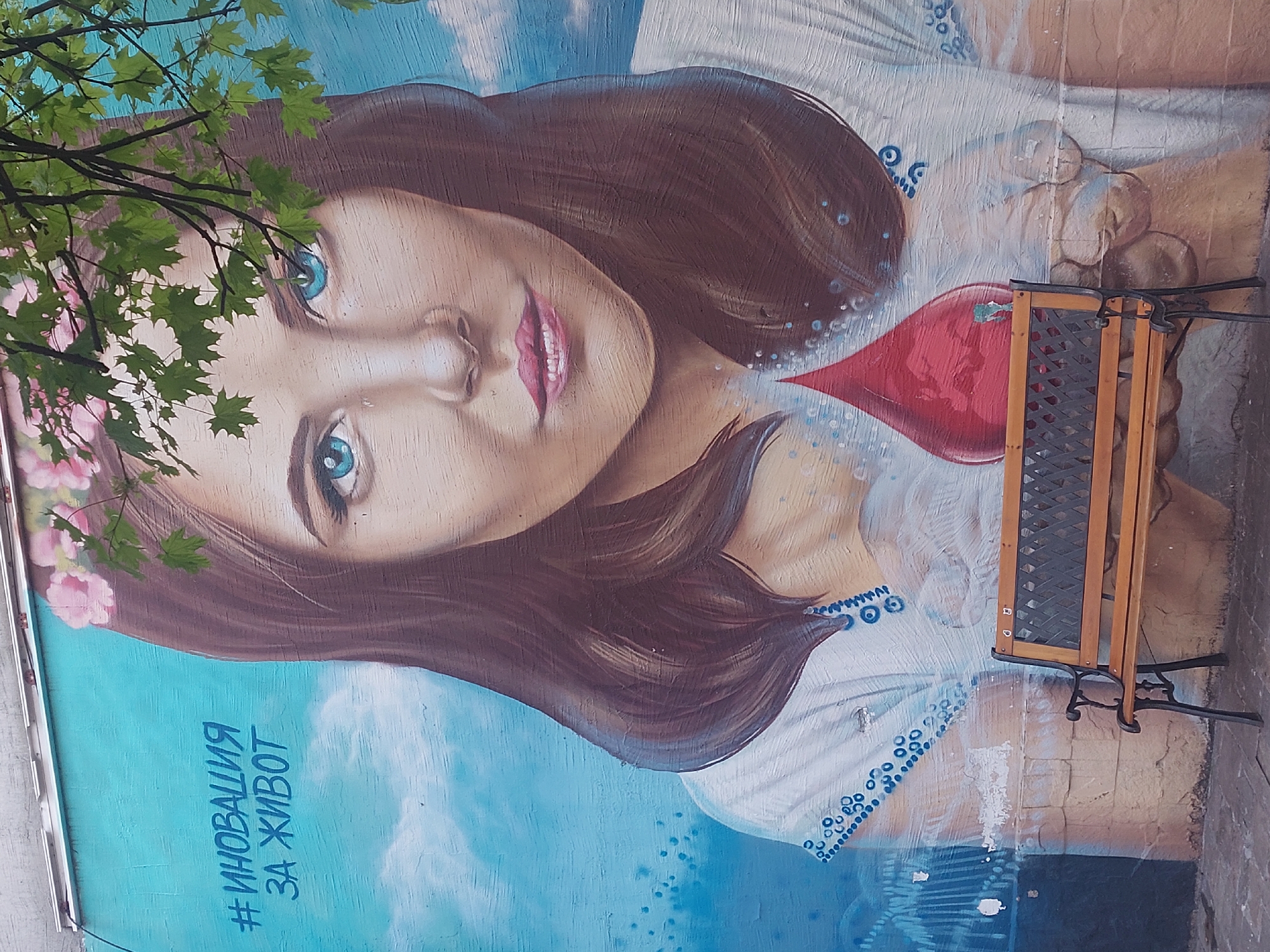 A mural depicting a woman with brown hair, blue eyes, and a flower crown on her head is visible. She is wearing a white shirt with blue embroidery. A red heart-shaped object is painted near her chest. The background consists of blue and white hues resembling clouds. The hashtag "#Иновация за живот" is painted in blue near the bottom left corner. A small wooden bench with a metal frame is situated in front of the mural on the right side of the image. A tree branch with green leaves is partially obscuring the upper left corner of the mural. The mural appears to be painted on a wall in Unknown.