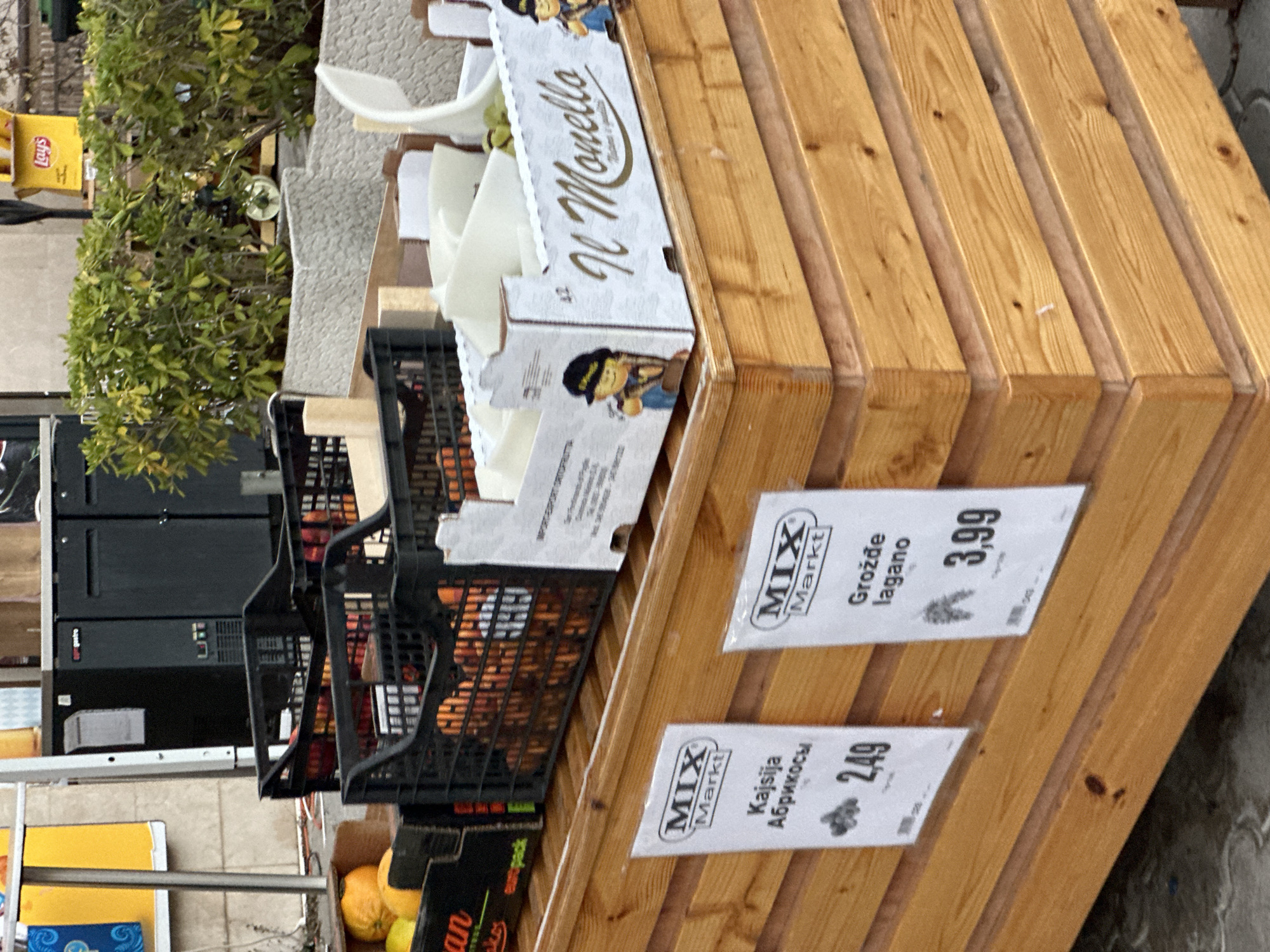 The image shows a market stall in Budva, Montenegro, with an assortment of fruits displayed in black crates and white boxes. In the foreground, two white signs with the "MIX Markt" logo are visible. The left sign indicates "Kajsija / Абрикосы" (Apricots) priced at 2,49. The right sign reads "Grožđe lagano" (Light grapes) priced at 3,99. Behind the signs, wooden shelves hold the produce. Apricots are visible in black crates, and a white box labeled "Il Monello" contains what appear to be pale fruits or vegetables, possibly melon or zucchini. Further back, black refrigerators or freezers are present, and lush green foliage is visible in the background. The stall appears to be outdoors, likely on a paved surface.