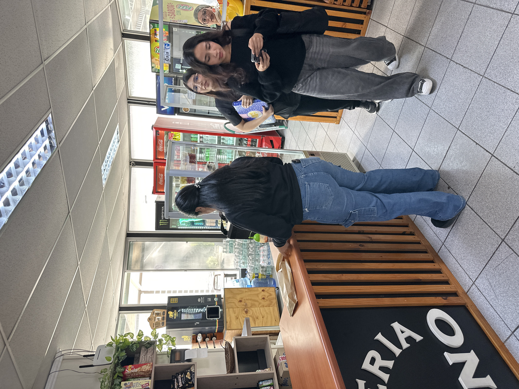 The image shows the interior of a small convenience store or "Oxxo" in Arica, Chile. At the checkout counter, a woman with long black hair in a ponytail wearing jeans and a black jacket is interacting with the cashier. Two other people are standing in the aisle. One woman is looking at her phone, the other is carrying a package of food. Behind them are refrigerated beverage coolers and a display of snacks. The interior has tiled floors and ceiling. The sign on the front of the counter appears to say "Riao" or "Oriaon." Shelves with various products are located behind the counter. Bottled water can be seen through the window behind the counter.