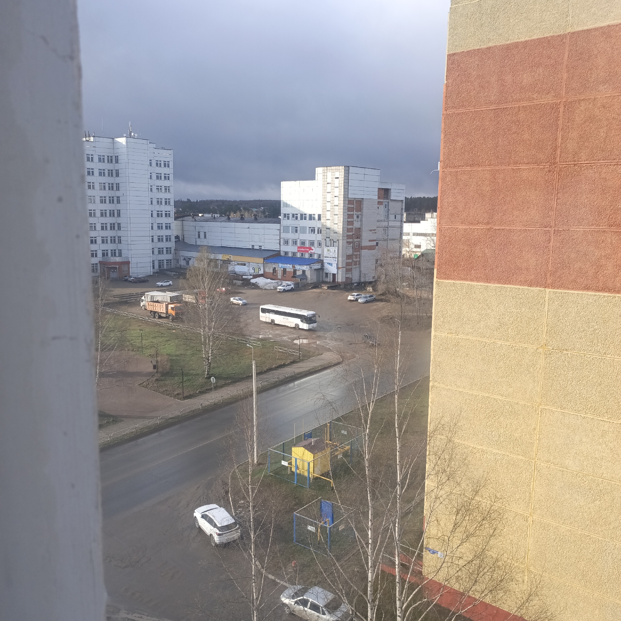 View from a high vantage point, likely a window, of a city street on an overcast day. In the foreground, parts of the building from which the photo is taken are visible: a white column on the left and a textured wall with blocks colored in shades of orange and yellow on the right. A street runs through the middle ground, and beyond it, there is a square with several parked cars and a bus. Two large white buildings dominate the background. A gas utility station is visible between bare trees, near the road, along with 2 cars. Bare trees are scattered throughout the scene. The sky is overcast.