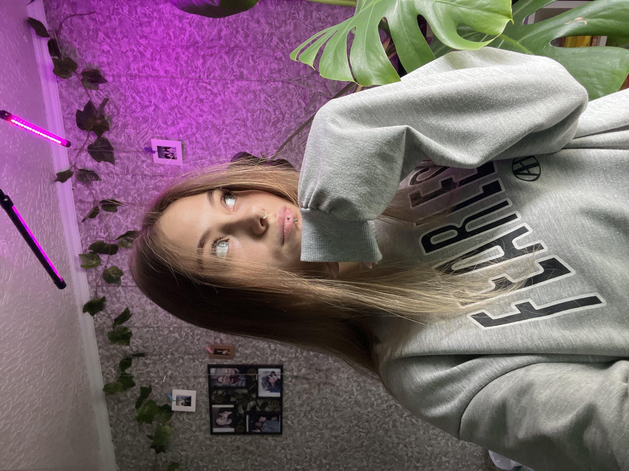 A young woman with blonde hair, nose ring, and lip piercings is facing upwards and towards the camera. She is wearing a grey hoodie with large black and white lettering. The background consists of a textured grey wall, a purple light source, green vine-like plants along the wall, a monstera plant in the upper right corner, and a framed collage of photos on the wall. The location cannot be determined as being in Фоки, Russia based on the provided image and available details.