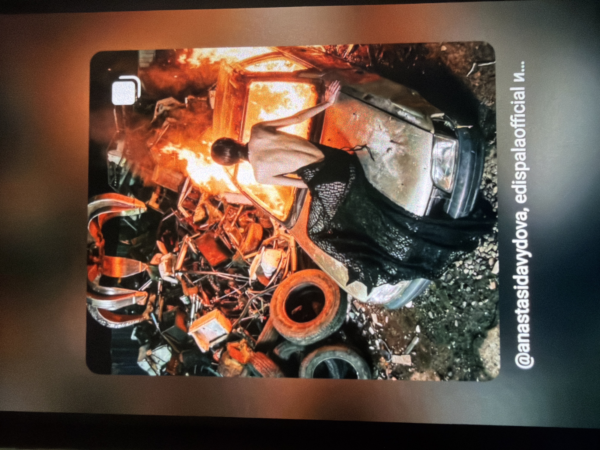 In a junkyard environment, a woman in a black, open-back dress sits on the hood of a burning car. The car is positioned amidst a pile of scrap metal, including various car parts and tires. Above the scene, a large clawed grapple hangs suspended, poised over the wreckage. The background is obscured by the fire and debris, but appears to be an industrial or demolition site. The overall impression is one of destruction and artistic posing. The image is framed by a digital interface, showing social media handles "@anastasidavyadova, edispalaofficial и...". There is no discernible information that would place this scene in Sofia, Bulgaria.
