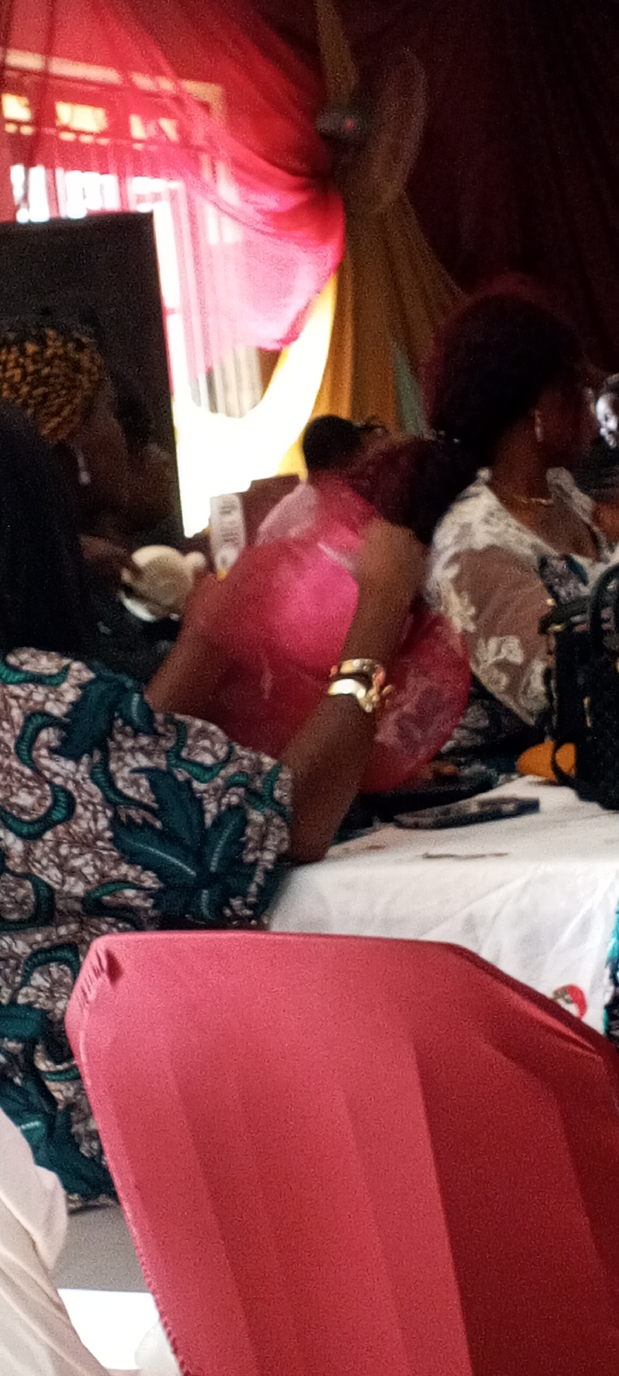 The image shows an indoor event with several people sitting at a table covered with a white cloth. One person is wearing a green and brown patterned dress, another is wearing a pink dress, and a third is wearing a white lacy top. A gold bracelet is visible on the arm of the person in the pink dress. The room is decorated with red and yellow drapery, and there appears to be a fan in the background. A red chair is in the foreground, partially obscuring the view of the table.