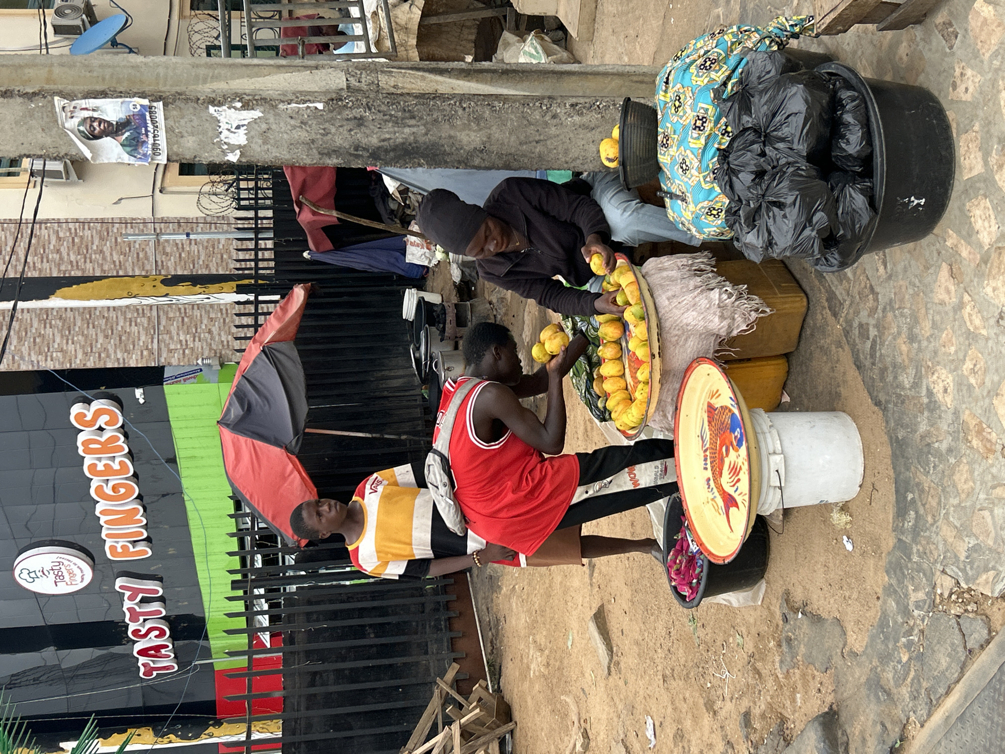 Two men sell fruit on the street in Jos, Nigeria. One man wears a black top and a black head covering; the other wears a red basketball jersey with black leggings and a white backpack. They are crouched beside a metal tray full of yellow fruits, seemingly mangoes or similar, and appear to be sorting the fruit. The fruit sits atop a white bucket, which is sitting on a black bucket, which is sitting on the sandy ground. Another black bucket full of black trash bags covered with colorful fabric sits to the right. A red and black umbrella is in the background. The men are in front of a restaurant called Tasty Fingers.
