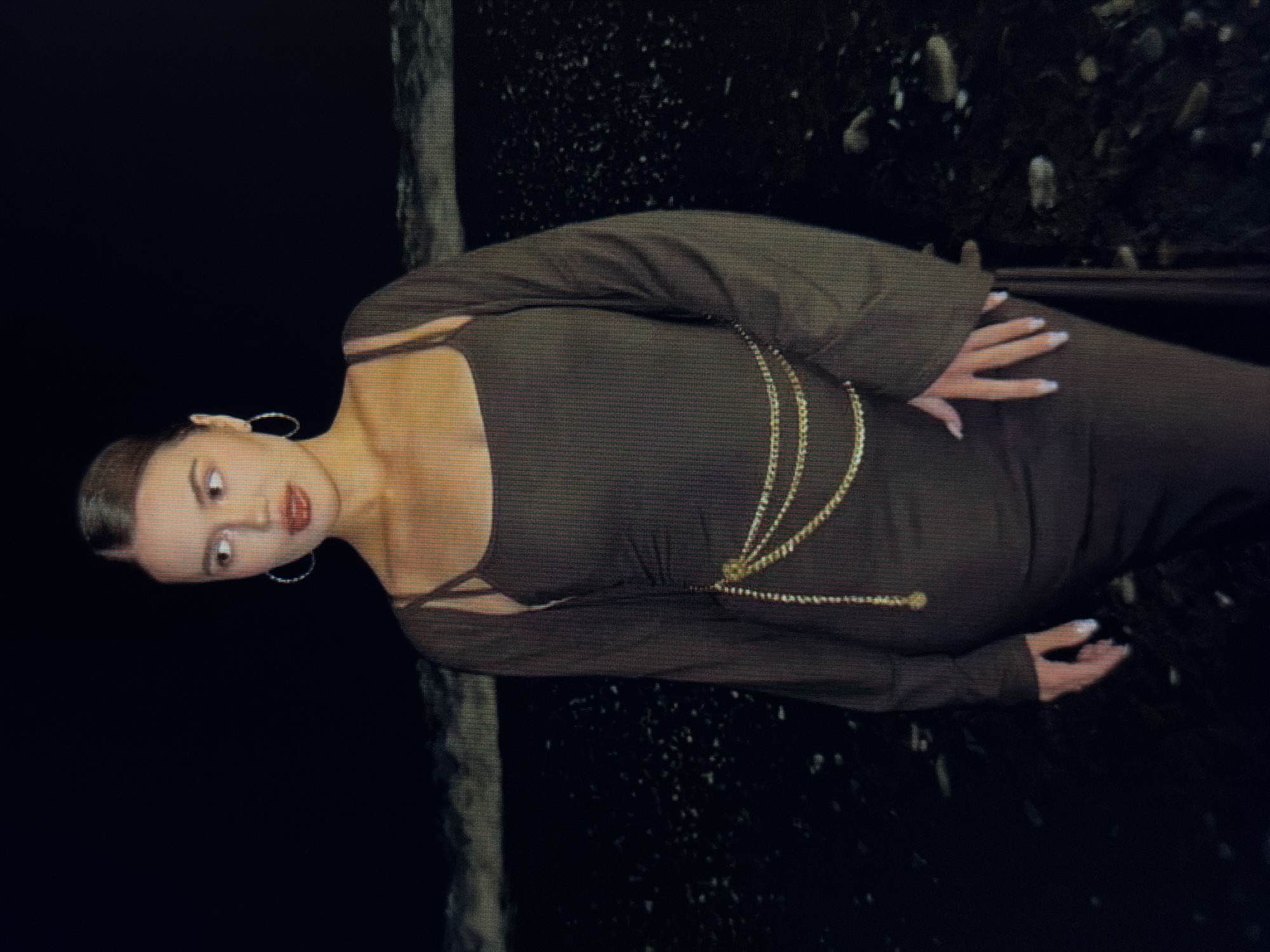 A young woman stands on a dark beach at night, facing away from the camera and looking to her right. She wears a dark, fitted dress with thin straps and a long-sleeved, open cardigan over it. A gold chain belt is cinched around her waist. Her hair is pulled back into a sleek bun. The background consists of a dark, choppy ocean with waves breaking on a dark, pebbly shore. The scene is dimly lit, with the woman's face and upper body illuminated by an unseen light source. The location is identified as Sirius, Russia.