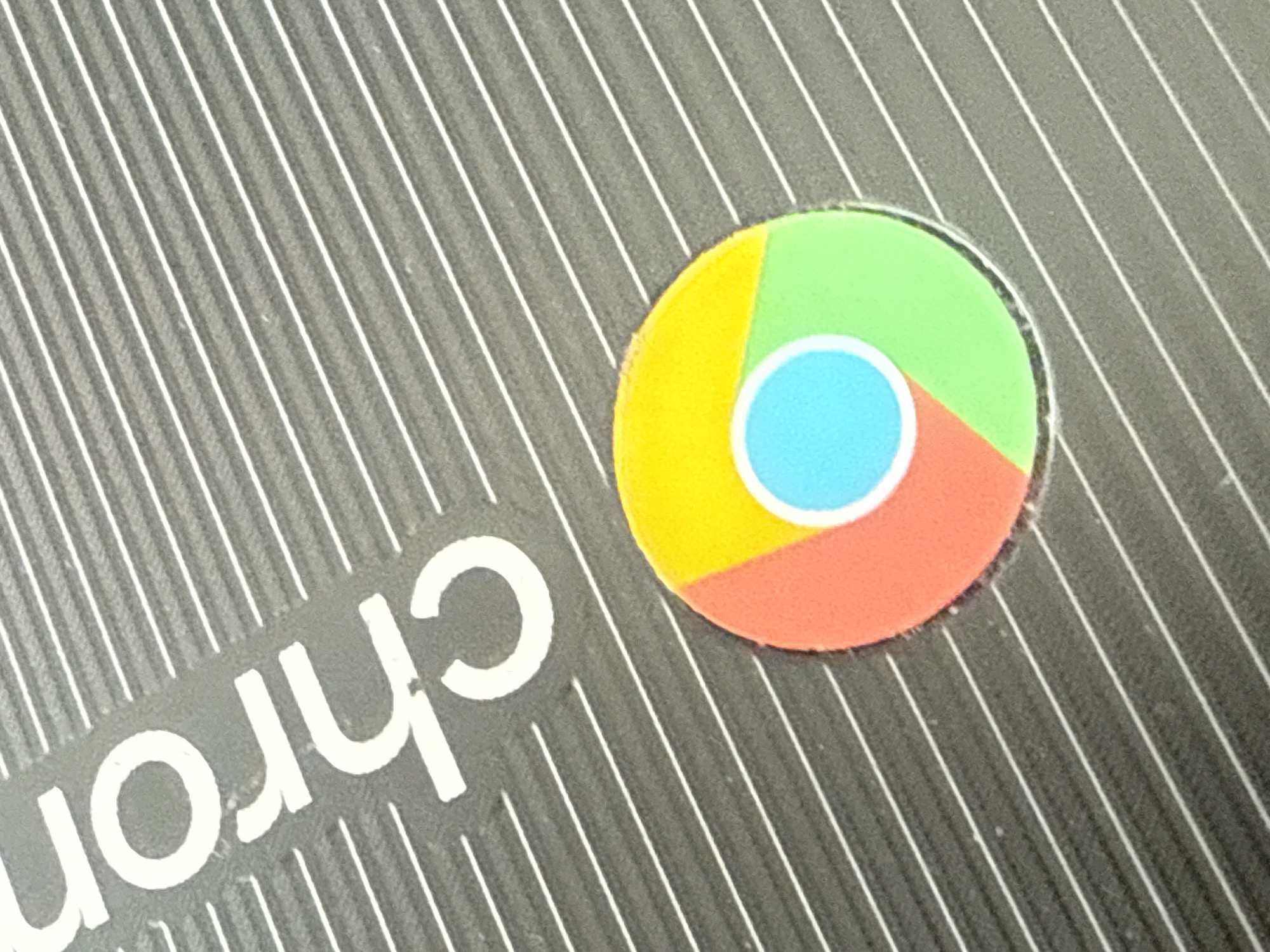 The image shows a close-up of a Chromebook laptop. The Chromebook logo, a colorful circular icon divided into green, yellow, and red sections with a blue circle in the center, is visible. The "Chrom" portion of the "Chromebook" branding is also partially visible, in white lettering on the dark grey laptop surface. The laptop surface is textured with parallel, diagonal white lines. The location is not discernible from the visual information.