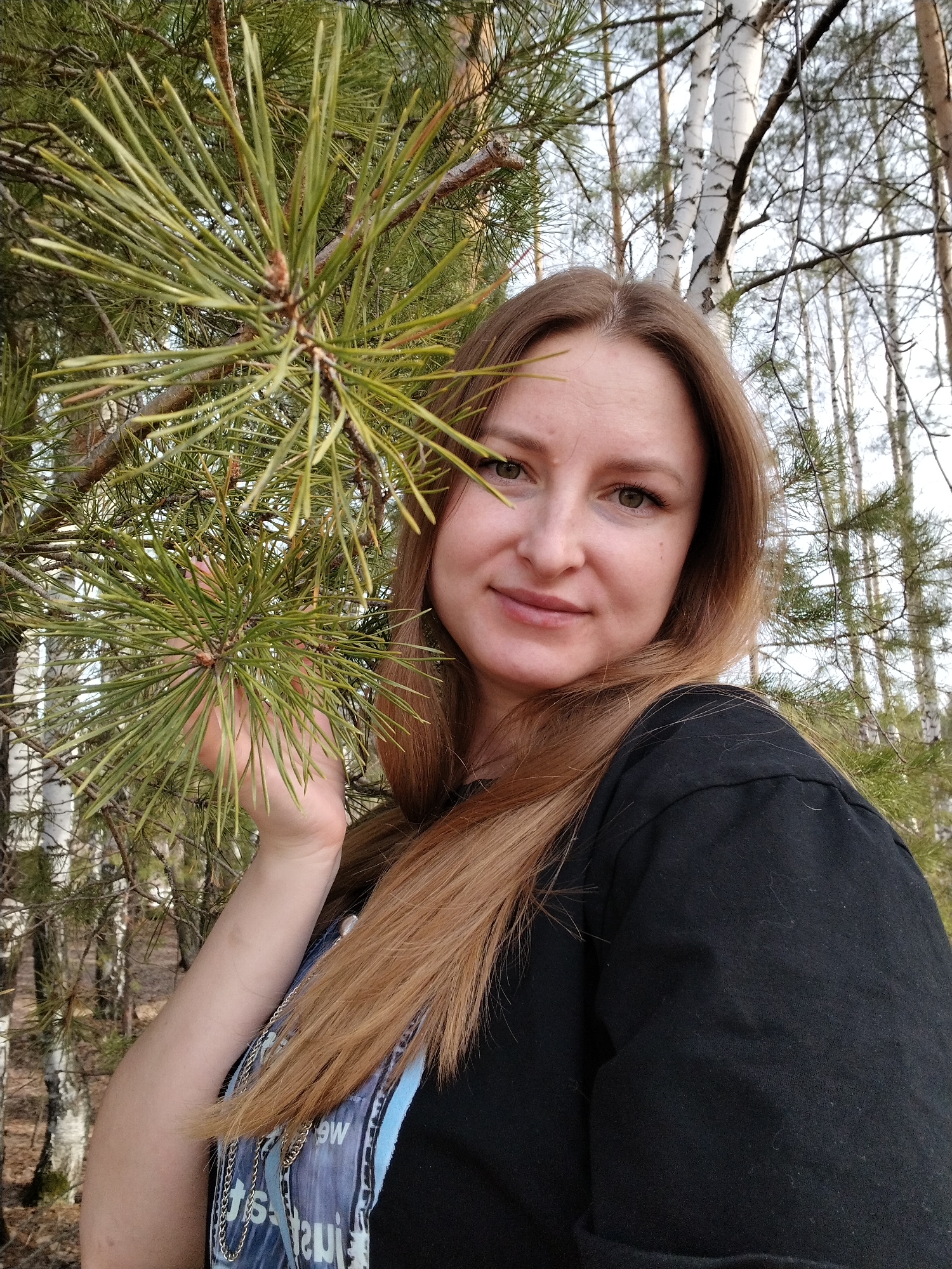A medium-angle outdoor shot features a fair-skinned woman with long, straight, reddish-brown hair, in what appears to be a forest. Her hair is parted to the side and falls over her right shoulder. She wears a black t-shirt, with the collar visible. Her left arm is extended, with her hand gently touching the needles of a green pine branch. The background consists of birch trees with white bark and thinner deciduous trees, indicating early spring or late autumn. The location is Chaikovsky, Russia.