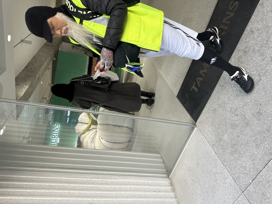 The image captures the entrance of a Tamburins retail store, likely located in Garosu-gil, Gangnam-gu, Seoul, South Korea, indicated by the "TAMBURINS" branded floor mat at the threshold.
In the foreground, a male subject with a long white beard and a black knit cap stands, dressed in a black puffer jacket over which a neon yellow high-visibility vest, marked with "SEOUL TOUR" vertically on the right strap, is worn. He also wears white Adidas athletic shorts, black compression socks, and black athletic shoes. His left forearm displays tattoos, and he holds a white object, possibly a phone, in his left hand. A black Nike fanny pack containing blue/green framed sunglasses is worn across his chest.
Reflected in the large glass entrance door/wall, a female subject with long dark hair, wearing a dark long coat with a fur collar, black boots, and carrying a black crossbody bag, is seen walking away from the viewer into the store's interior. Visible inside the store through the glass is a large, segmented white pumpkin-like art installation with a golden stem. The reflection also prominently displays the text "BURINS".
The exterior features light grey speckled flooring, leading to the black "TAMBURINS" floor mat. A textured, vertically ribbed light grey wall forms the left boundary of the entrance. The interior beyond the reflected female shows a green wall further back and recessed lighting overhead.