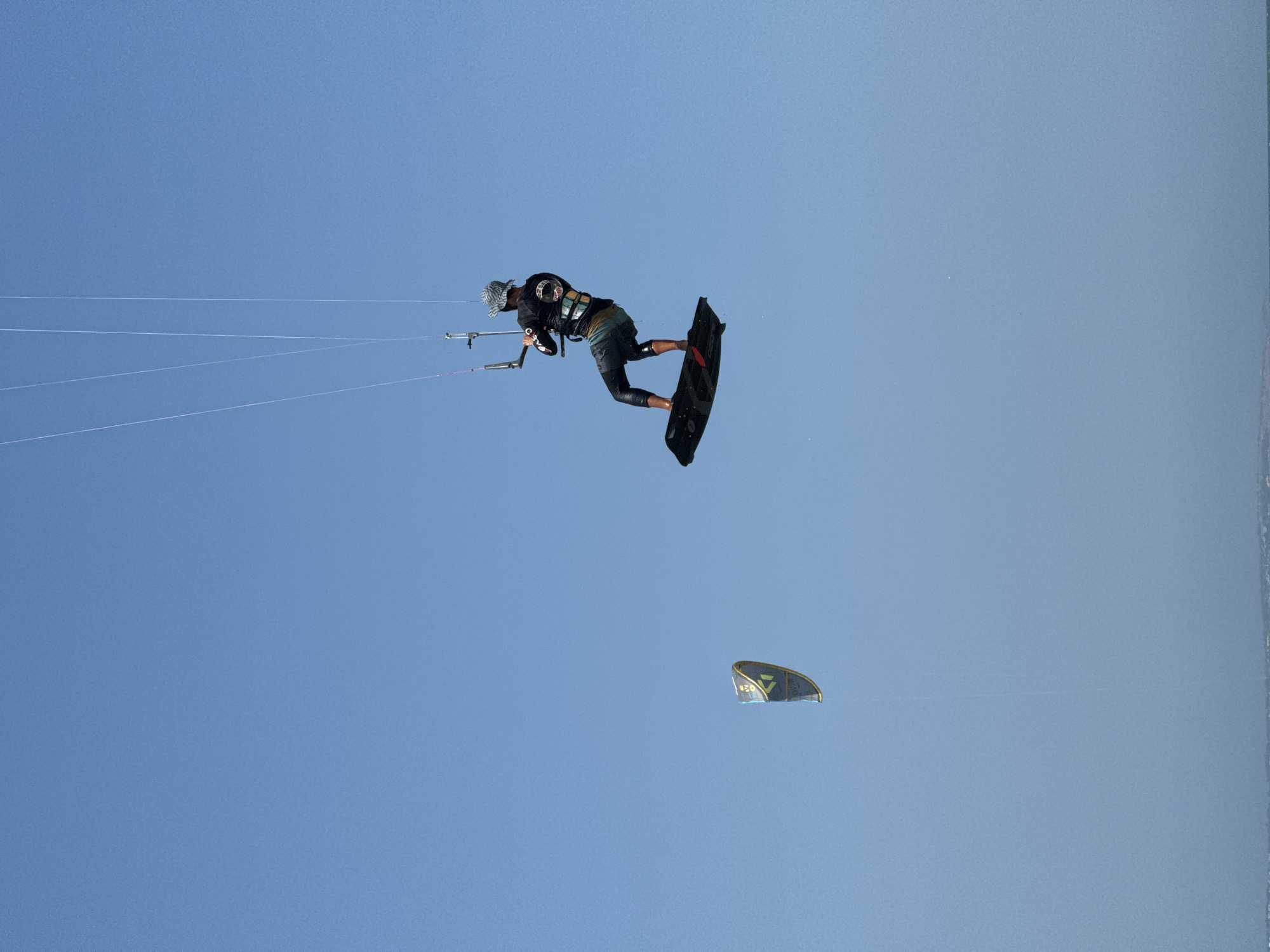 A kitesurfer is captured mid-air, suspended against a clear blue sky. The kitesurfer, wearing a black long-sleeved shirt, dark shorts, and a patterned hat, is leaning back on a black board, holding onto a kite bar connected to a kite. Another kite, with a yellow and blue design, is visible in the distance to the left. The image is taken from below, looking up at the kitesurfers and the sky. The bottom edge of the image shows a faint line of what appears to be land or distant objects, suggesting the kitesurfing is taking place over water, likely near the coast of Mũi Né, Vietnam. The lighting suggests it is daytime.