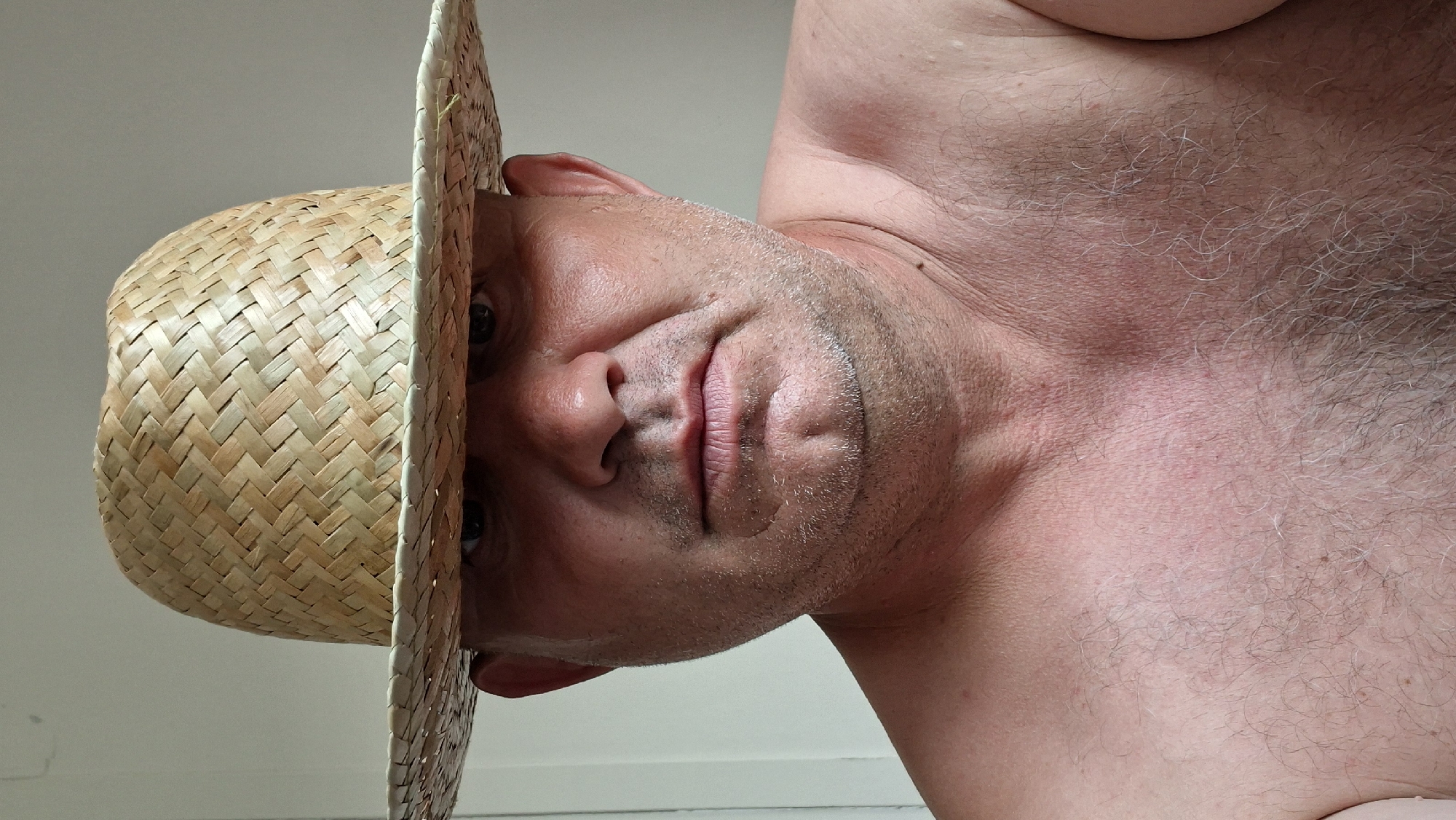 A shirtless middle-aged man with fair skin and stubble is wearing a woven straw hat that casts a shadow over his eyes. He is looking directly at the camera with a neutral expression. The background is a plain, light-colored wall. The image appears to be a selfie taken indoors, possibly in Hîncești, Moldova, given the request's context. His chest and upper shoulders are visible and appear to be lightly tanned with some sparse, graying body hair.