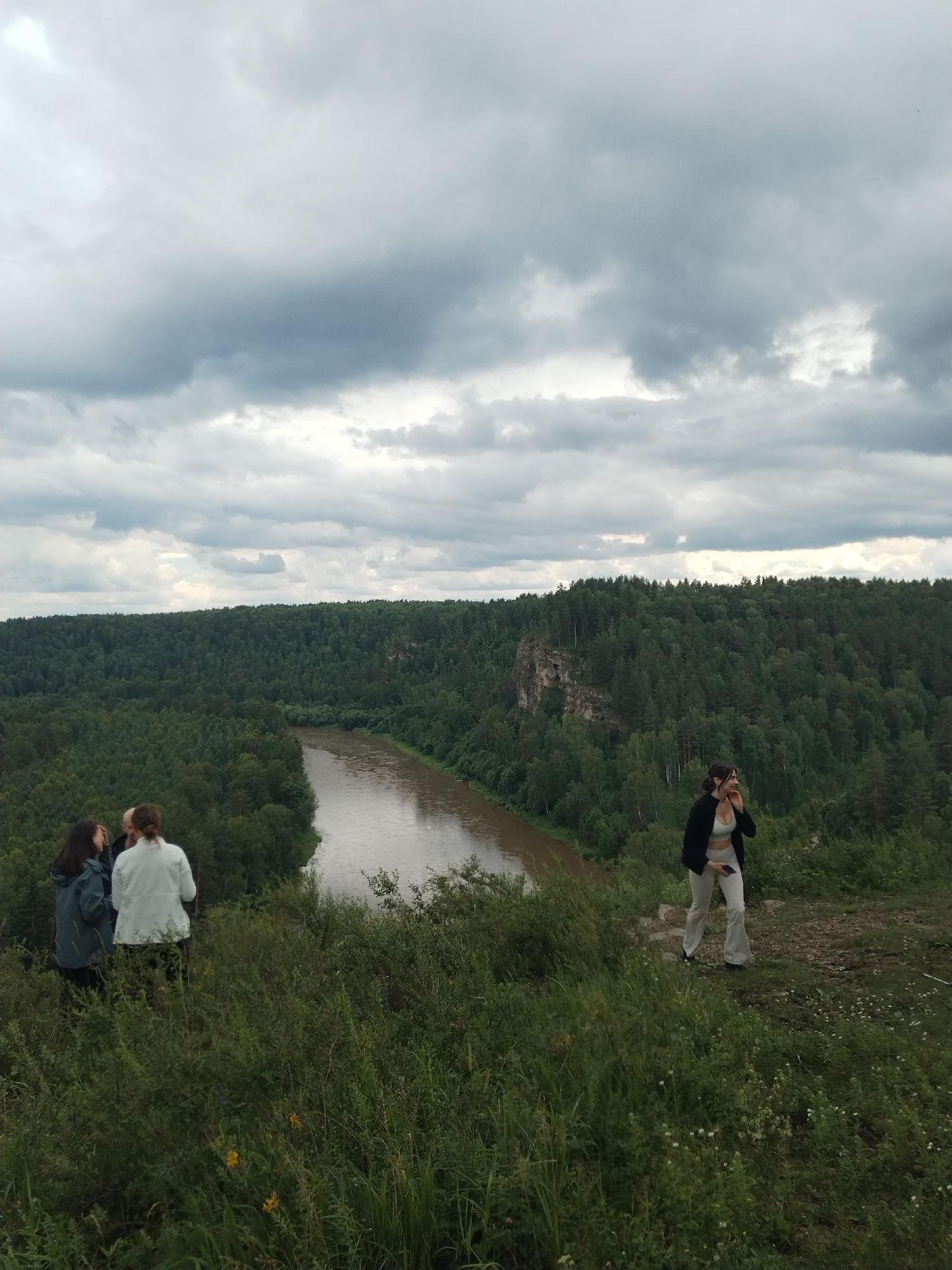 This image depicts a scenic overlook in Idrisovo, Russia, featuring a winding river bordered by dense, green forest. The sky is overcast with dramatic clouds. On the left side of the frame, three individuals are gathered on a grassy knoll, their backs mostly to the camera. One person is wearing a dark jacket, another a light-colored jacket, and the third's attire is less distinct. To the right, a woman stands on a dirt path, wearing a white crop top and light-colored flared pants, with a dark jacket draped over her shoulders. She is looking away from the camera and appears to be holding a phone. The foreground is filled with tall, green grass and some small white and yellow flowers, suggesting a natural, undeveloped area. The overall scene conveys a sense of exploration and appreciation for the natural landscape.