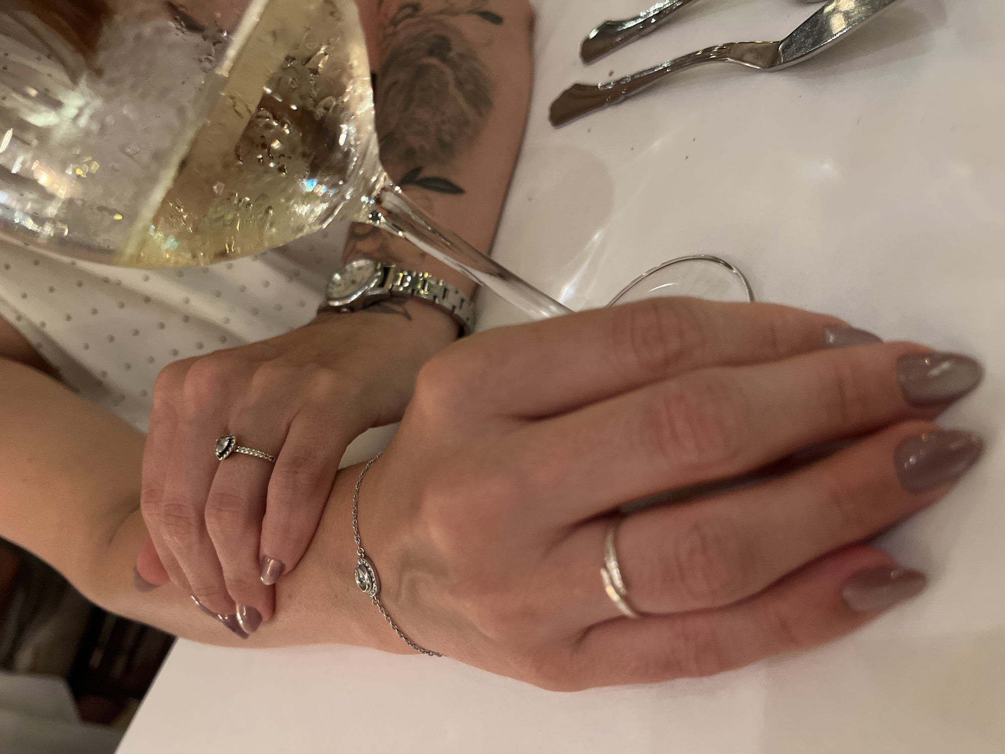 A close-up, waist-up shot shows a woman's hands and arms resting on a white table. She is wearing a white dress with a subtle heart pattern and a silver watch. On her left wrist, a delicate silver bracelet with a clear circular pendant is visible. Her left hand is crossed over her right arm, adorned with a silver ring featuring a dark, possibly black, teardrop-shaped stone. Her right hand, resting on the table, wears a thin silver band. Her fingernails are painted in a taupe color. A wine glass filled with a pale yellow liquid, likely white wine or champagne, is held by her right hand, with condensation visible on the glass. The background is slightly blurred, hinting at an indoor setting, possibly a restaurant. In the bottom right corner, a silver fork and knife are partially visible. The image is taken from a slightly high angle. There is no specific indication of the location being Barcelona, Spain.