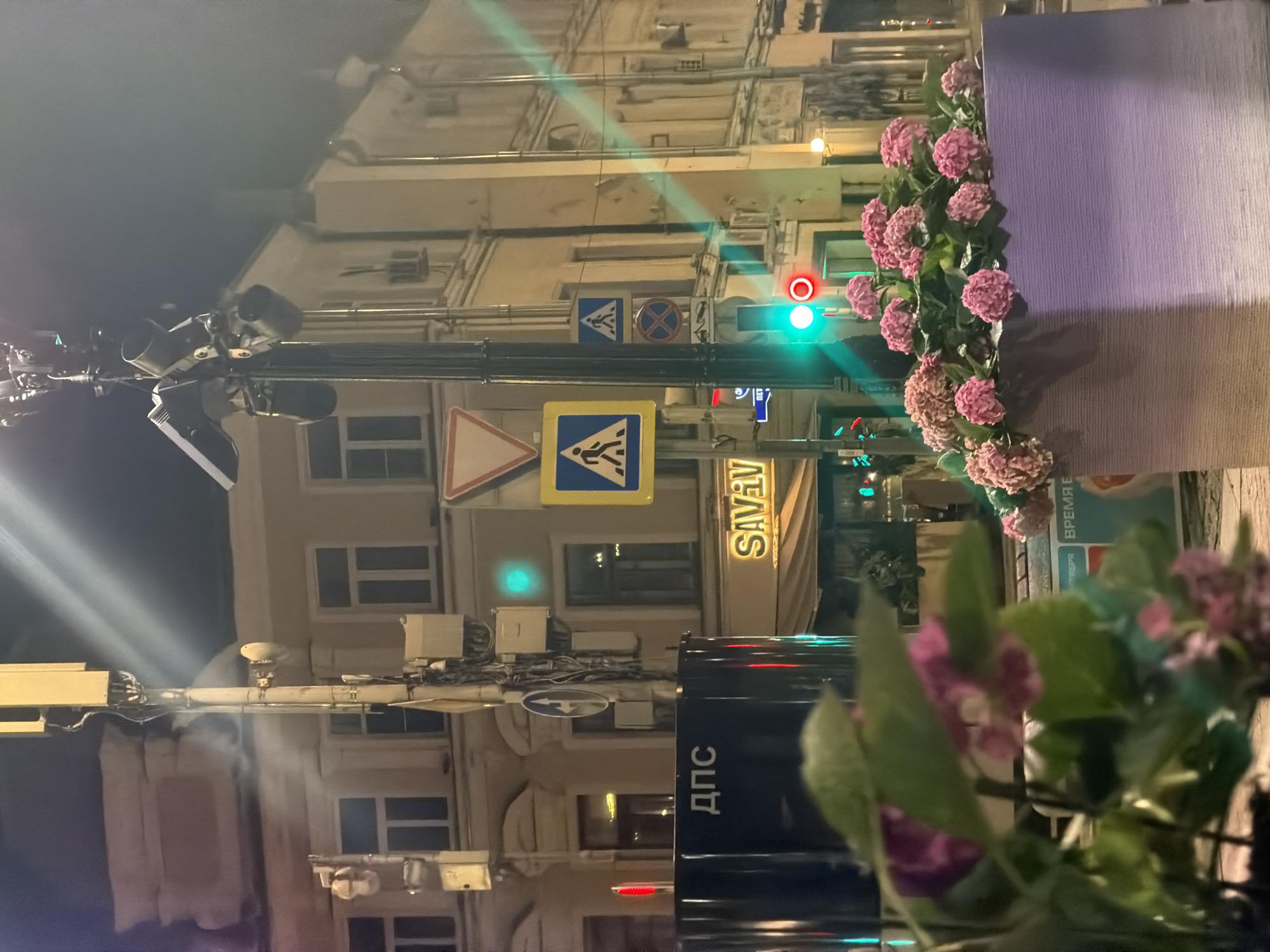 The image depicts a nighttime street scene in Moscow, Russia. In the foreground, a planter box filled with pink hydrangeas is visible. Behind the planter, a dark blue vehicle with white Cyrillic lettering "ДПС" (DPS, traffic police) is parked. To the right of the vehicle, a building with windows and a sign that reads "SAVANNA" is illuminated. Various traffic lights and street signs are present, including pedestrian crossing signs and a "no stopping" sign. A tall pole with multiple antennas and lights is a prominent feature in the background, suggesting a busy urban environment. The lighting suggests it is nighttime, with streetlights and building lights illuminating the scene. The overall impression is that of a bustling city street at night.