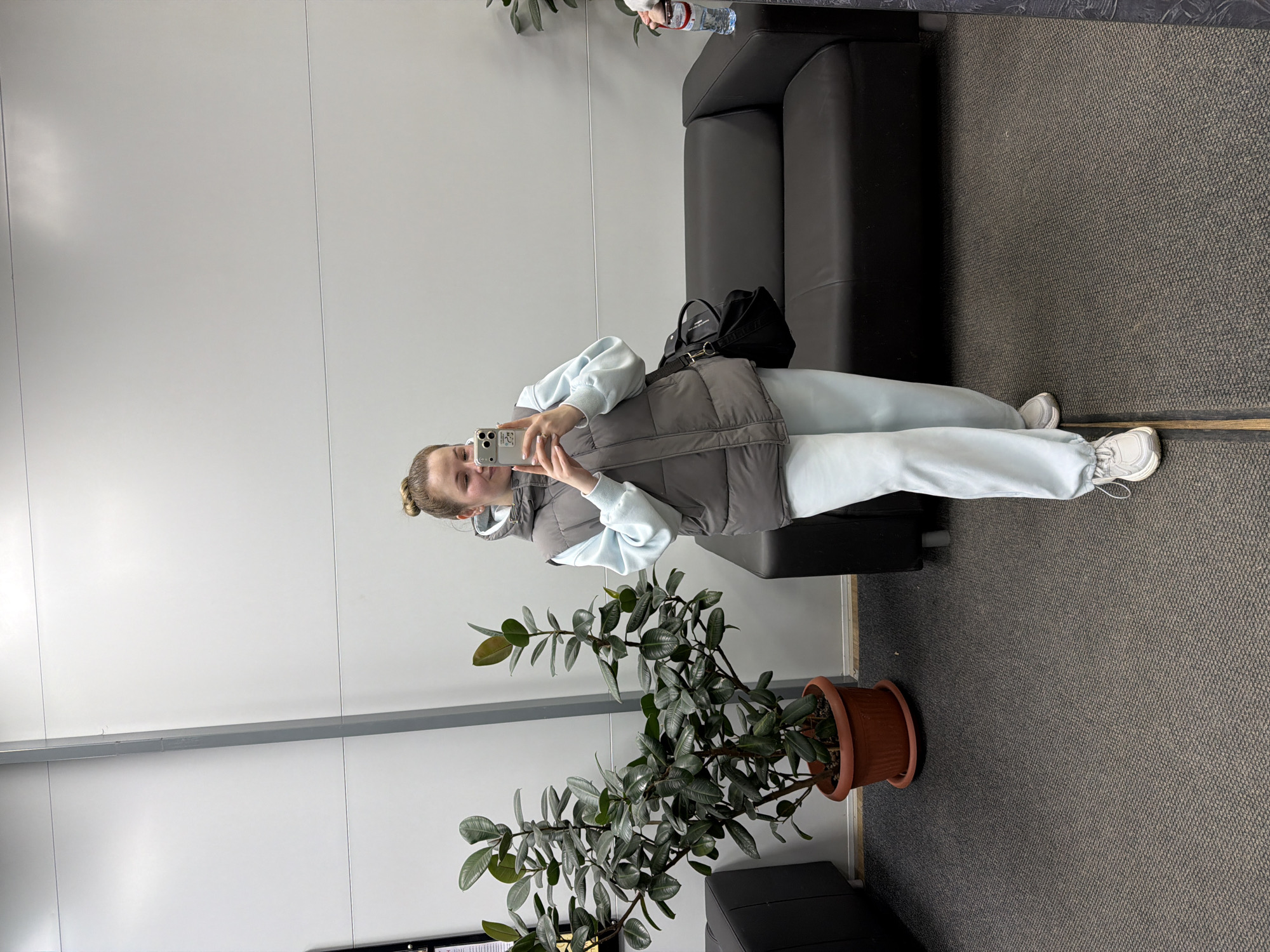 A young woman is captured in a mirror selfie indoors, positioned slightly to the right of the frame. She holds a smartphone in both hands, looking towards it with a neutral expression and a faint smile. Her light-colored hair is pulled back into a bun.
She is dressed casually in a light blue long-sleeved top, matching light blue sweatpants, a grey puffer vest, and white sneakers. Behind her, a black handbag rests on a black leather sofa.
The background features plain white paneled walls, with a horizontal grey bar or shelf dividing them across the mid-section. The floor is covered with a grey carpet. In the foreground, to the left, a large potted plant with dark green leaves stands on the carpet. On the armrest of the sofa, a plastic water bottle is visible, with the brand name "BON AQUA" clearly readable on its label. The lighting appears bright and even, indicating an indoor environment that resembles a public or commercial waiting area or lounge.