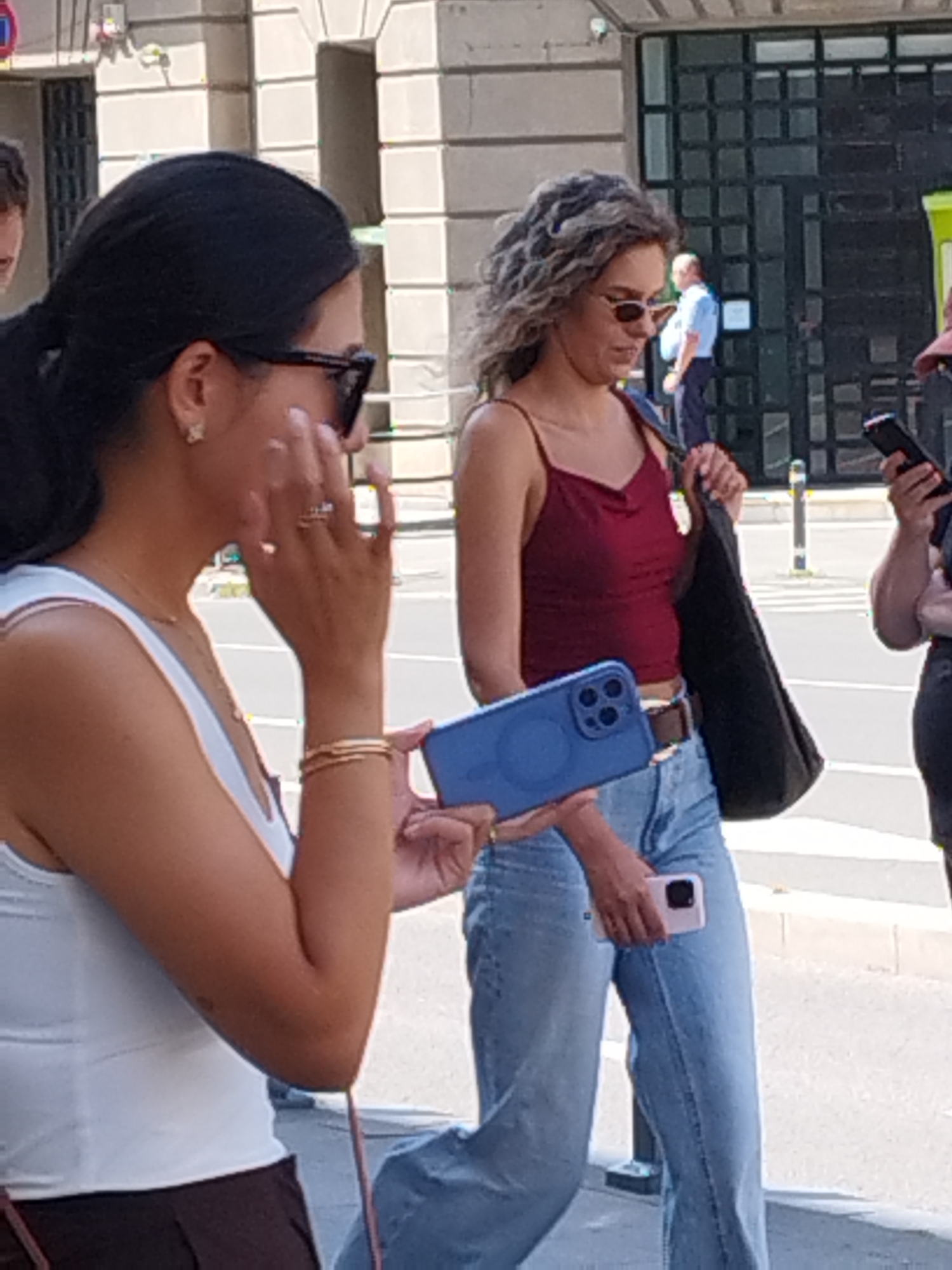 Two women are walking on a street in Bucharest, Romania. The woman on the left is wearing a white tank top and dark pants, with her hair pulled back. She is holding a blue phone with a circular attachment in her right hand. The woman on the right has curly hair and is wearing a burgundy tank top and light blue jeans. She is carrying a black tote bag. In the background, a man in a blue shirt and dark pants is standing near a building. The street appears to be paved with cobblestones, and there are planters with greenery visible.