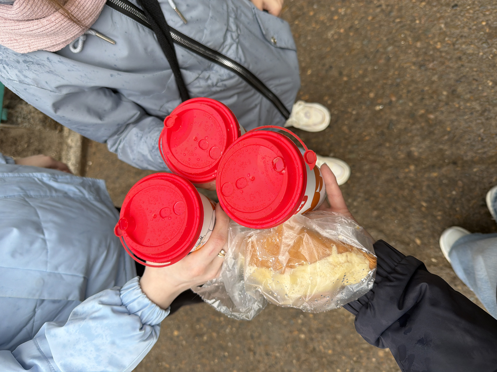 Three individuals are shown from an overhead perspective, each holding an item. One person, wearing a light blue jacket, holds a coffee cup with a red lid. A second individual, in a light grey puffer jacket and a pink knit hat, also has a coffee cup with a red lid. The third person, dressed in a dark blue jacket, is holding a plastic bag containing a sandwich or pastry. The ground beneath the people appears to be a paved surface, possibly a street or sidewalk. The location is Chaikovsky, Russia.