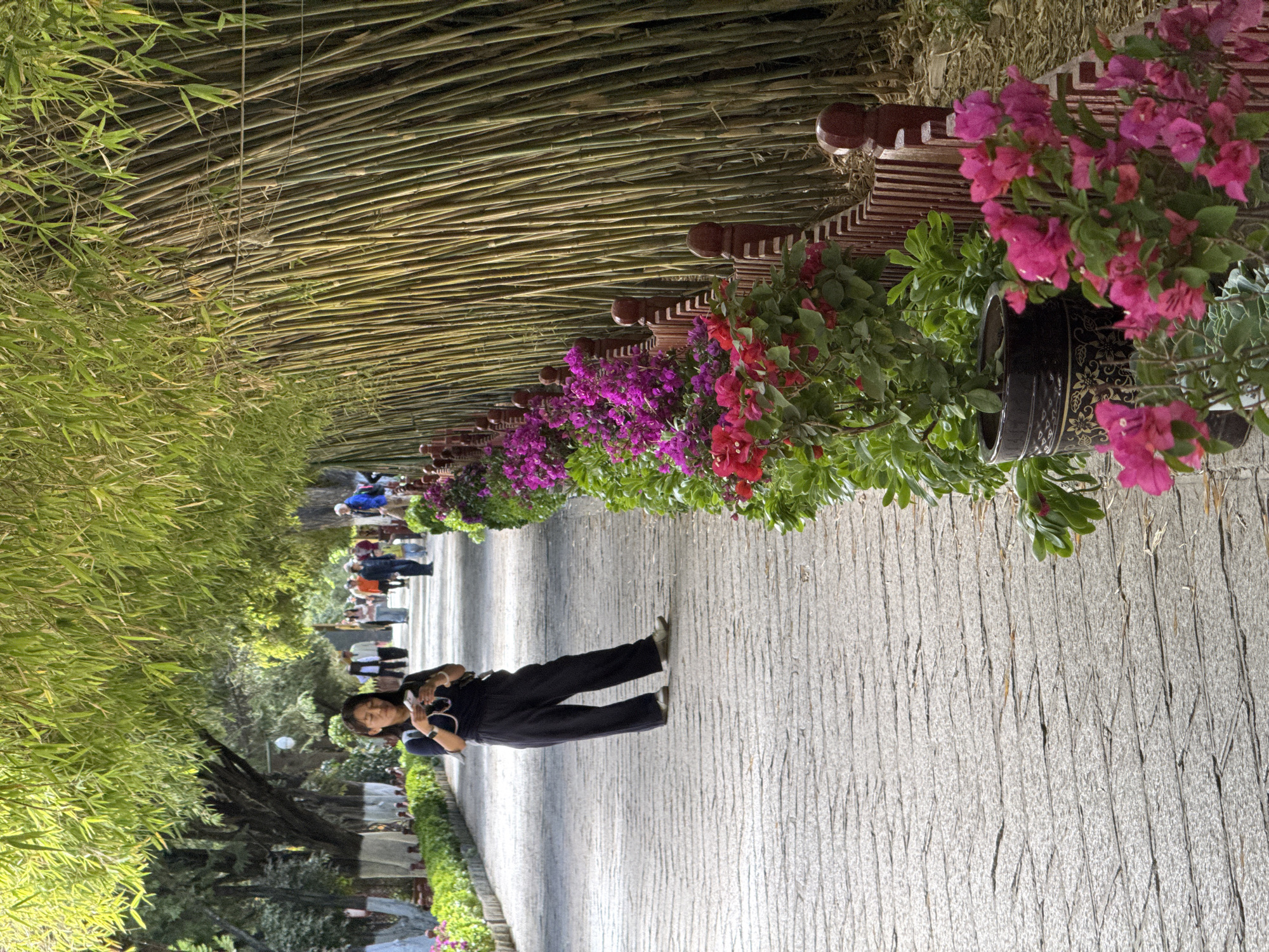 A horizontal, ground-level shot shows a tree and flower-lined walkway covered by a bamboo roof. A woman wearing a navy blue shirt and navy blue pants stands on the walkway facing away from the camera and toward a group of people walking. The left of the walkway is lined with trees while the right is lined with pots containing flowers. The flowers are pink, purple, and red. The sidewalk is gray and has a linear pattern. Above the walkway, bamboo is spread horizontally to act as a roof. The location is in the city of Chengguan District, China.