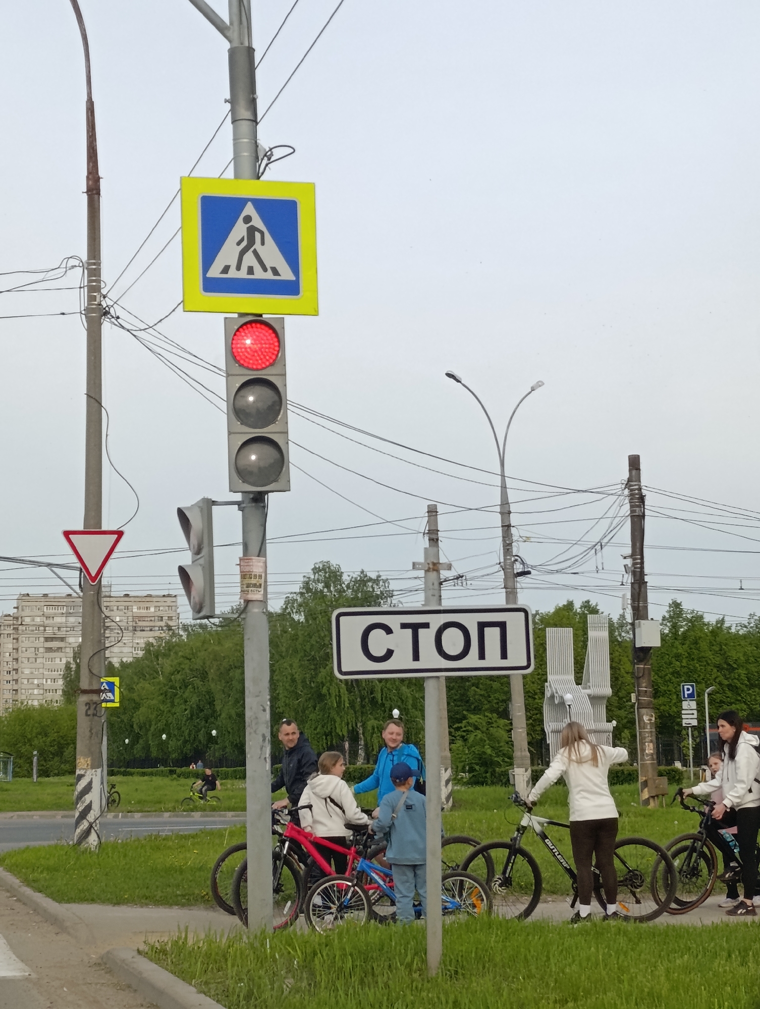 The image captures a city scene in an unknown location. A traffic signal pole dominates the foreground, adorned with a pedestrian crossing sign, a traffic light displaying red, and a "STOP" sign. A yield sign is attached to an adjacent pole.
A group of people, including adults and children, are gathered near the traffic signal with their bicycles. They appear to be stationary and possibly interacting. Trees are visible in the background, along with buildings suggesting a residential or urban area. Power lines crisscross the sky, and streetlights are also visible.