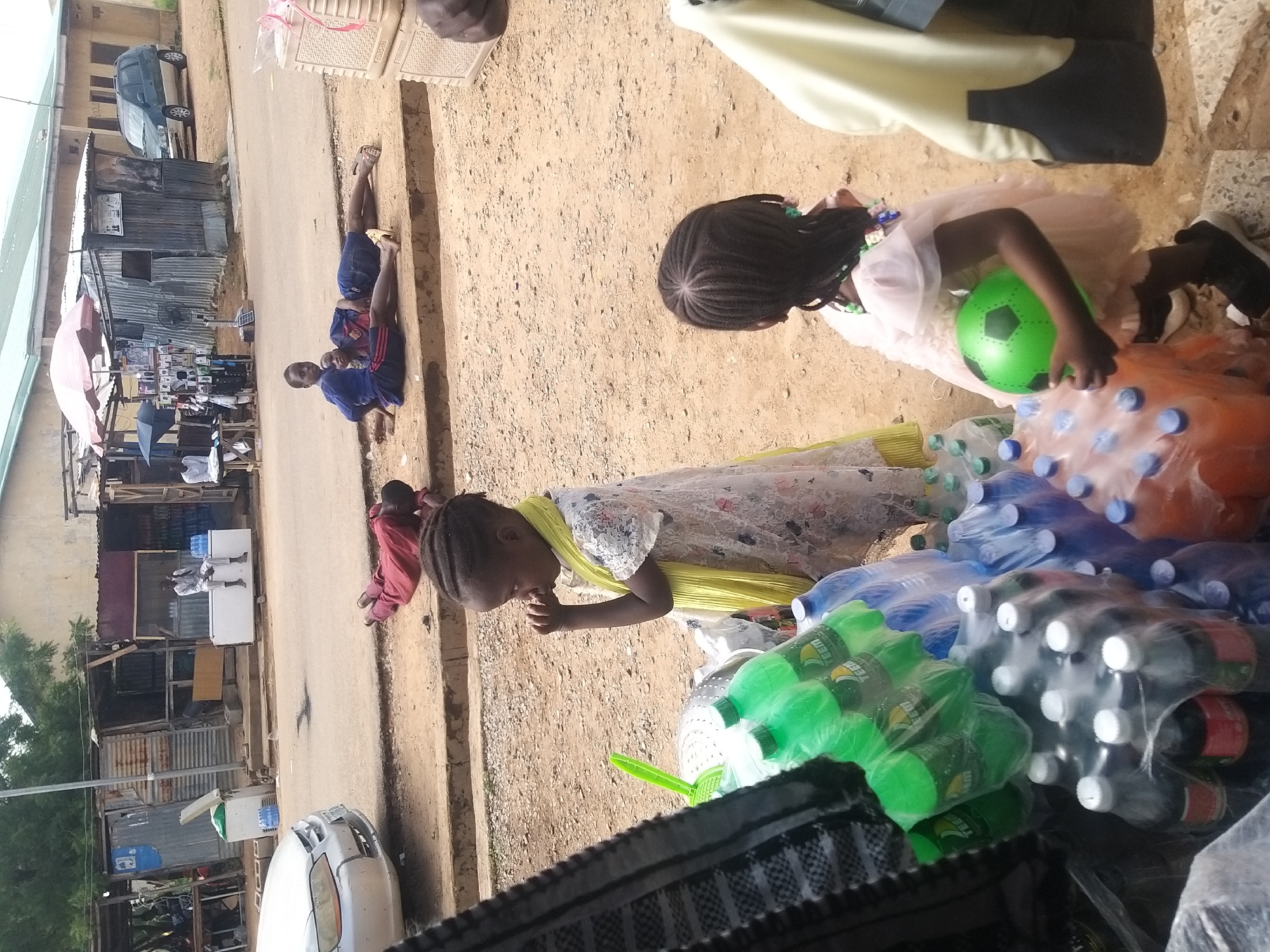 The image depicts a street scene in Kano, Nigeria, characterized by a dusty, unpaved surface and a row of small shops or stalls lining the street. In the foreground, two young girls are visible. One, in a white dress with a yellow scarf, stands near a display of bottled drinks, appearing to observe something. The other girl, in a pink dress, holds a green soccer ball and is facing away from the camera. Behind them, a collection of bottled beverages in plastic packaging is arranged.
Further back, on the street, a person in a red garment is lying face down on the ground. Two other individuals, appearing to be children, are lying down on the dirt shoulder of the road, one with a blue top and the other in a blue shirt and dark shorts. In the background, several shops with corrugated metal roofs and open fronts are visible. A light-colored car is parked on the street to the left, and a dark green SUV is parked further down the road to the right. People are seen interacting within or near the shops. The overall environment suggests a commercial area in a developing urban setting.