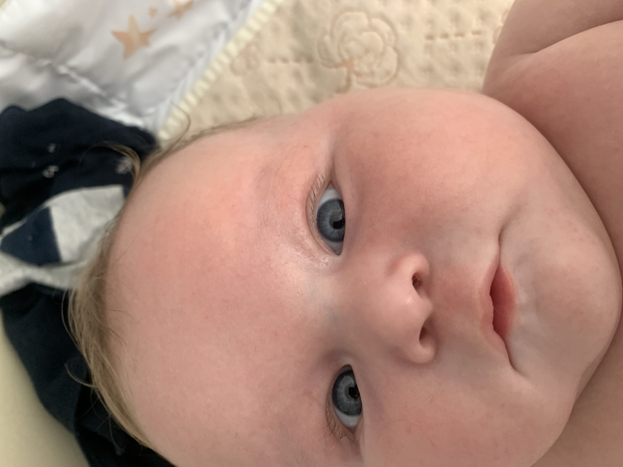 The media file is a close-up, overhead shot of a baby's face. The baby has fair skin, light blonde hair, and blue eyes. The baby is lying on its back on a soft, patterned surface, possibly a blanket or mattress. To the left of the baby's head, a dark blue fabric with a white stripe pattern is visible, suggesting clothing or bedding. The lighting appears to be natural, casting soft shadows. The baby's expression is neutral, with its mouth slightly open. The overall scene conveys a sense of domestic intimacy and care. No text is visible in the image. The location context is Stavropol, Russia.