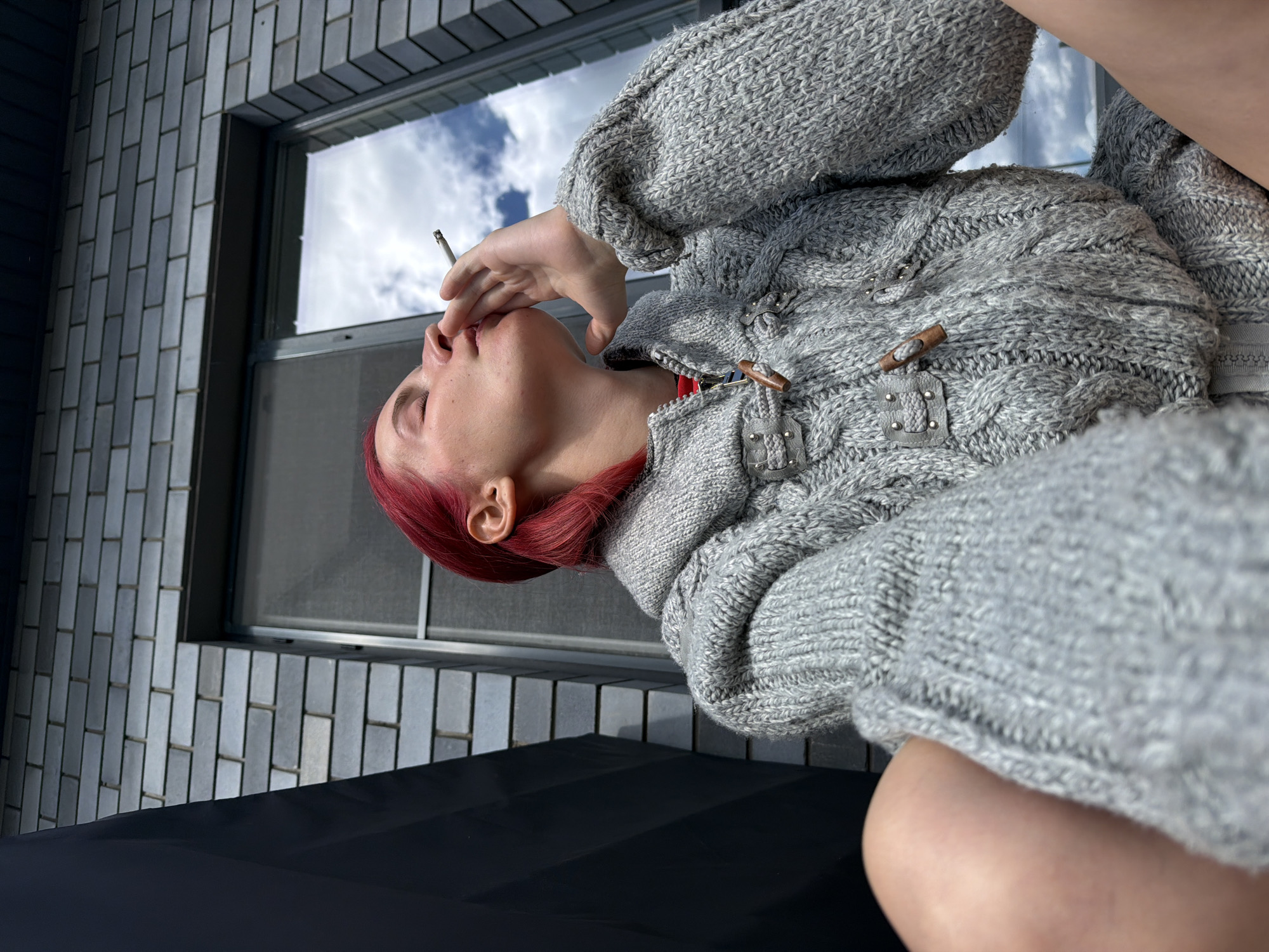 A person with bright red hair is sitting outside and smoking. They are wearing a thick, grey, cable-knit sweater with wooden toggle closures. The person's eyes are closed as they inhale from the cigarette, which is held between their fingers. In the background, a modern building with dark grey horizontal siding and a large window reflecting the sky is visible. The camera angle is low, looking up at the person and the building. The scene appears to be in an urban or suburban environment, possibly on a balcony or terrace.